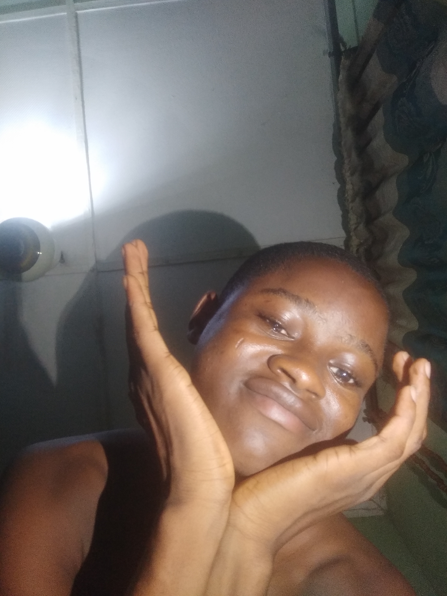 A close-up, indoor portrait features an individual with dark skin and short, dark hair, smiling gently at the camera. Their hands are positioned on either side of their face, cupping it softly. The person's expression is one of contentment or mild amusement, with a soft smile and slightly narrowed eyes. A subtle sheen is visible on their skin, particularly on the cheeks and forehead.
The background consists of a light-colored wall or ceiling, illuminated by a bright, artificial light source originating from the upper left corner of the frame. This light casts prominent, dark shadows of the individual's head and raised arm onto the wall behind them. A circular light fixture is partially visible in the upper left area. To the right, a textured or patterned fabric, possibly a curtain, can be seen. The individual's bare shoulders are visible, suggesting they may not be wearing a shirt. No text is visible in the image.