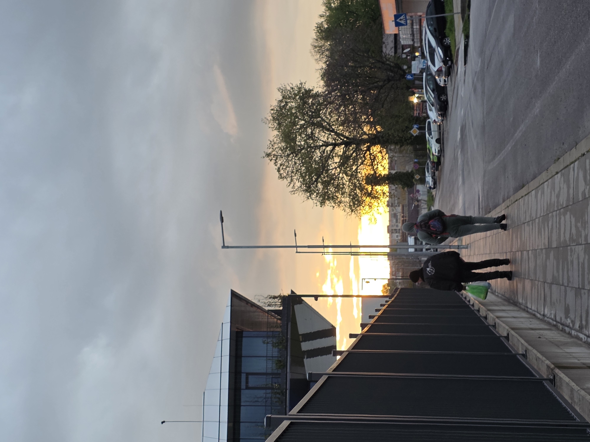 The image shows a cityscape at sunset in Plovdiv, Bulgaria. The scene is taken from an elevated perspective, likely a pedestrian bridge or walkway. Below, modern architecture with glass and steel facades is visible. Two pedestrians are walking along the walkway. One is wearing a dark jacket and carrying a green bag. The other is wearing lighter clothing and a backpack. A row of parked cars lines the street on the right side of the image. Street lights are visible. In the background, the setting sun creates a golden glow in the sky and illuminates the trees, adding a warm contrast to the cooler tones of the sky and the urban environment.