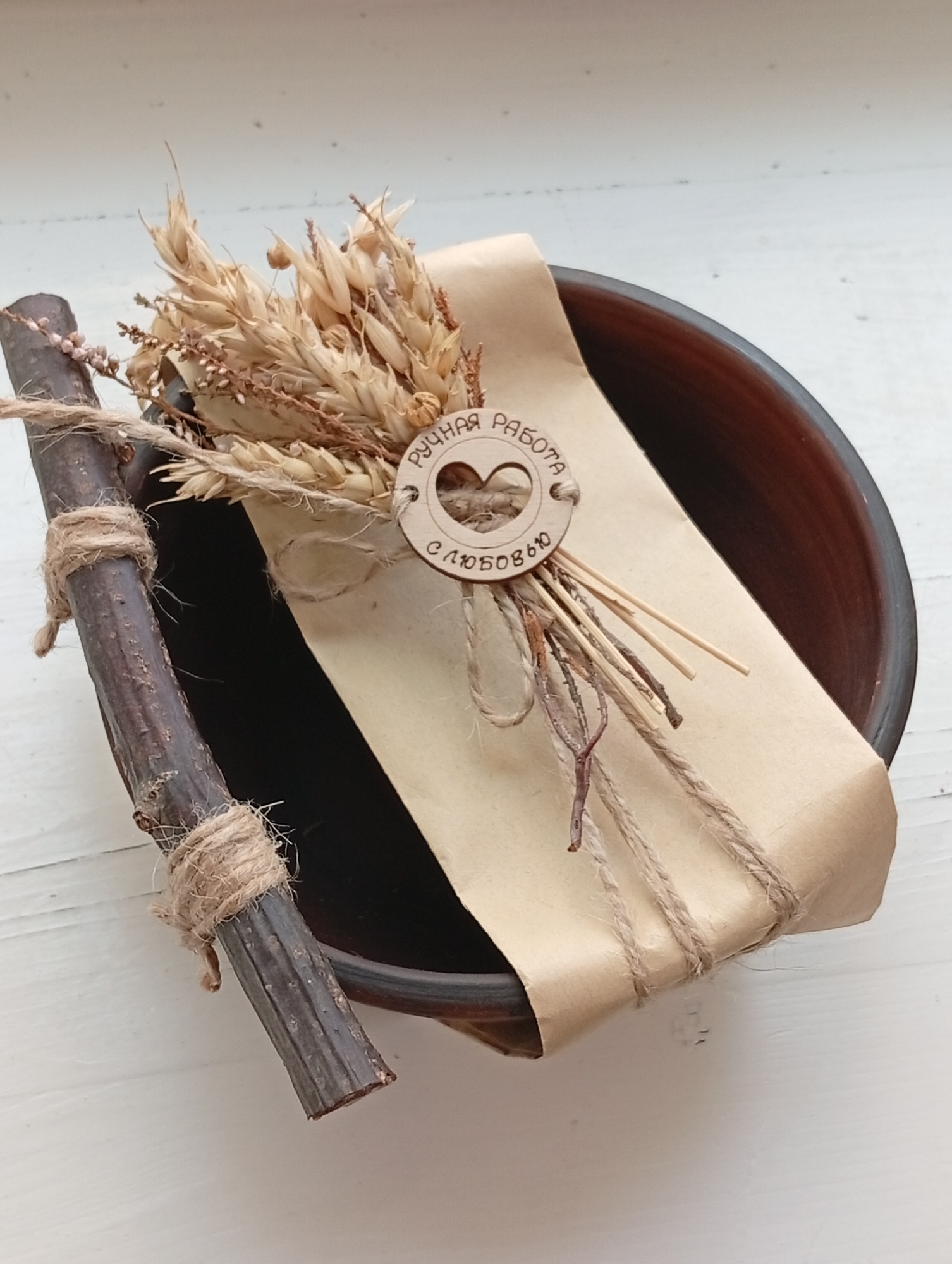A still life photograph features a rustic arrangement of natural elements. In the foreground, a dark, gnarled stick is tied with jute twine, lying diagonally across a textured, light-colored surface. Behind the stick, a dark brown ceramic bowl is visible, containing a bouquet of dried wheat stalks and small, dried flowers. The bouquet is wrapped in brown kraft paper, secured with jute twine. A small, circular wooden disc with carved text and a heart shape is attached to the wrapping, adding a decorative detail. The overall impression is one of natural, handmade craft. This scene does not depict any urban environment and therefore cannot be placed within the city of Крыўлянскі сельскі Савет, Belarus.