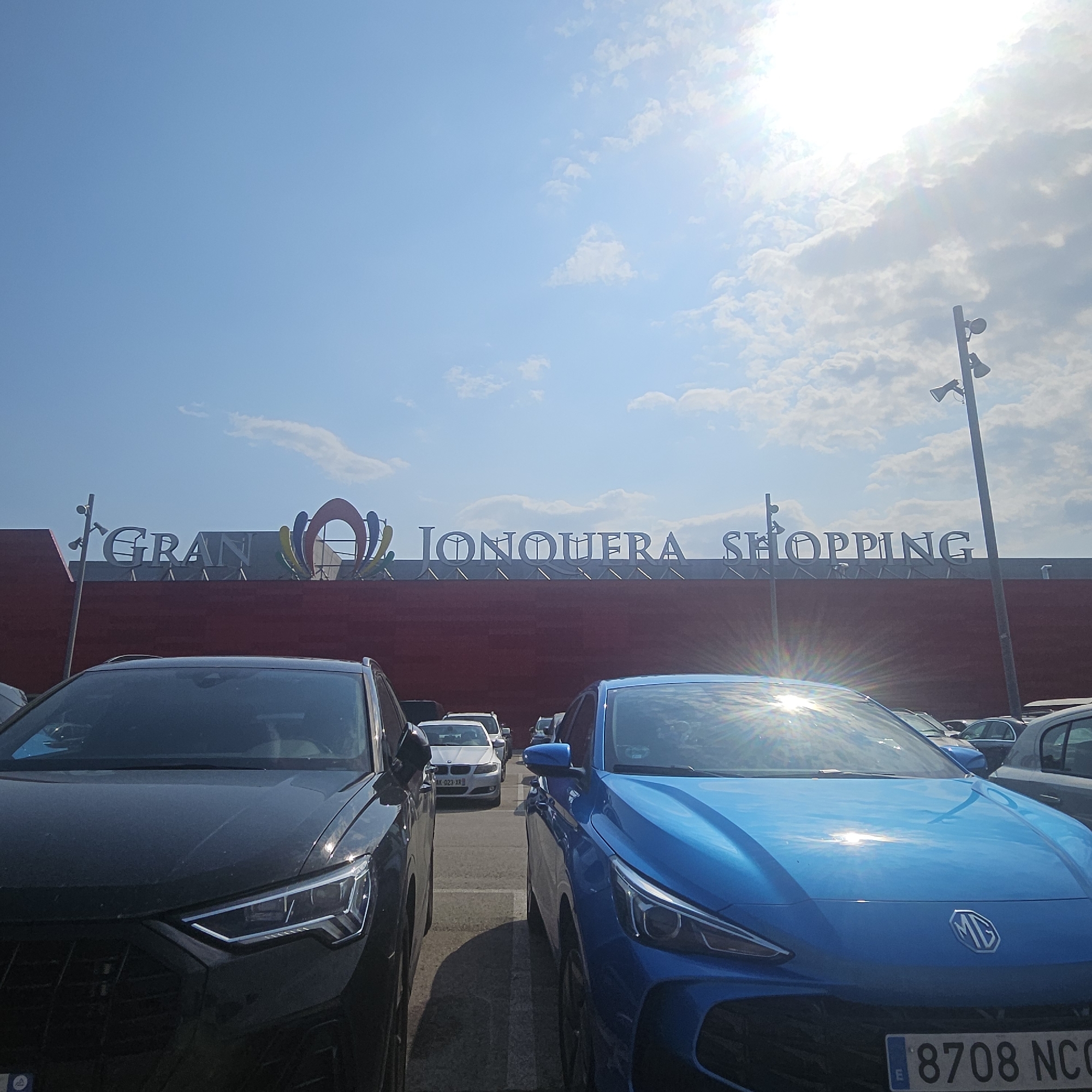 The image shows a view of the Gran Jonquera Shopping center in La Jonquera, Spain, under a bright, sunny sky with scattered clouds. In the foreground, two cars are parked, a dark-colored SUV on the left and a bright blue sedan on the right. The blue sedan has an MG logo on its grille and a license plate reading "8708 NCC". Behind the cars, the red facade of the shopping center is visible, with large white lettering that spells out "GRAN JONQUERA SHOPPING". The sky is a clear blue, with the sun positioned high and slightly to the right, causing lens flare and reflections on the cars. A tall pole with two speakers or lights is visible on the right side of the shopping center. In the background, other cars are parked in a lot, and the architecture of the shopping center suggests a commercial area. The overall scene depicts a typical daytime view of a shopping center in Spain.