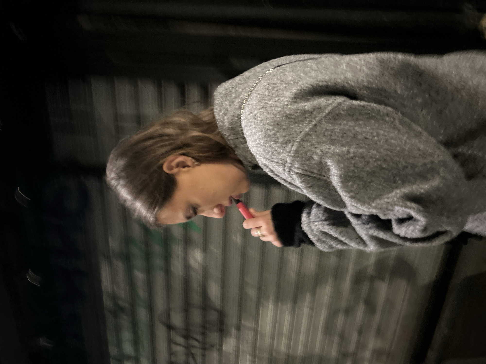 A person is holding a pink electronic cigarette device in front of a vertically corrugated metal wall at night. The person is wearing a gray coat or sweater over a black sleeve. They are standing or walking along a street, as evidenced by the dark, blurred area above the metal wall. The location is a city, though the specific city is unknown.