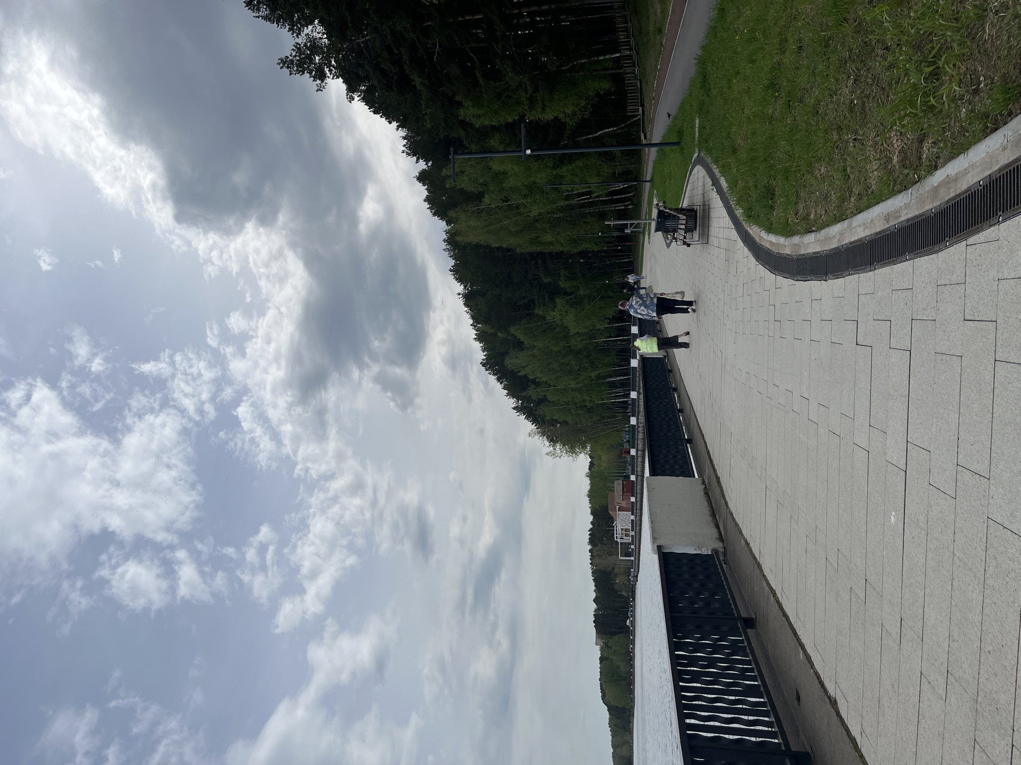 The image captures a view of a pedestrian walkway in the city of Chaikovsky, Perm Krai, Russia. The walkway, constructed of light grey paving stones, runs parallel to a dense forest. A black metal drainage system separates the walkway from a strip of grass along the forest's edge. There are two people standing on the walkway; one is wearing a light green vest. The sky is overcast with cloudy conditions. Metal lamp posts are positioned along the walkway. The image is taken at an angle, appearing rotated.