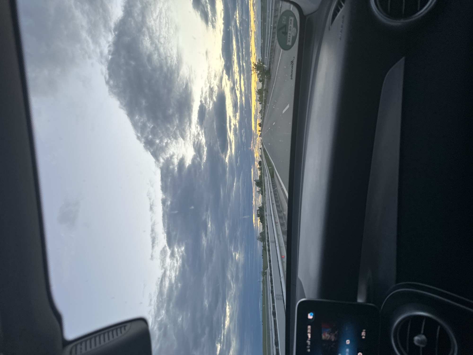 The image, taken from inside a car, shows a view of a highway in the city of Dobri Dol, Bulgaria. The road stretches into the distance under a sky filled with clouds. The car's dashboard and air vents are visible in the foreground. A small screen in the vehicle is also visible.