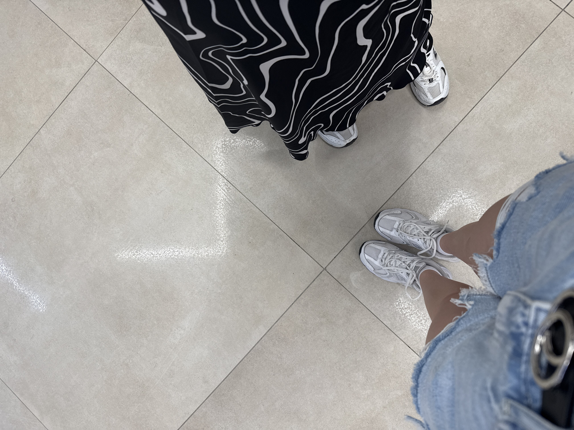 This is a first-person perspective, overhead shot of two individuals standing on a light-colored tiled floor. One person is wearing a long black and white patterned dress and white sneakers. The other person is wearing denim shorts and white sneakers. The setting appears to be indoors, possibly a retail environment or a hallway, given the clean, uniform tiling. The lighting suggests it's daytime, but there are no clear indicators of the time of day or weather outside. The focus is on the lower half of the individuals, specifically their footwear and lower garments. There are no visible emotions or specific activity beyond standing. No text is visible in the image. The location context is Chișinău, Moldova.