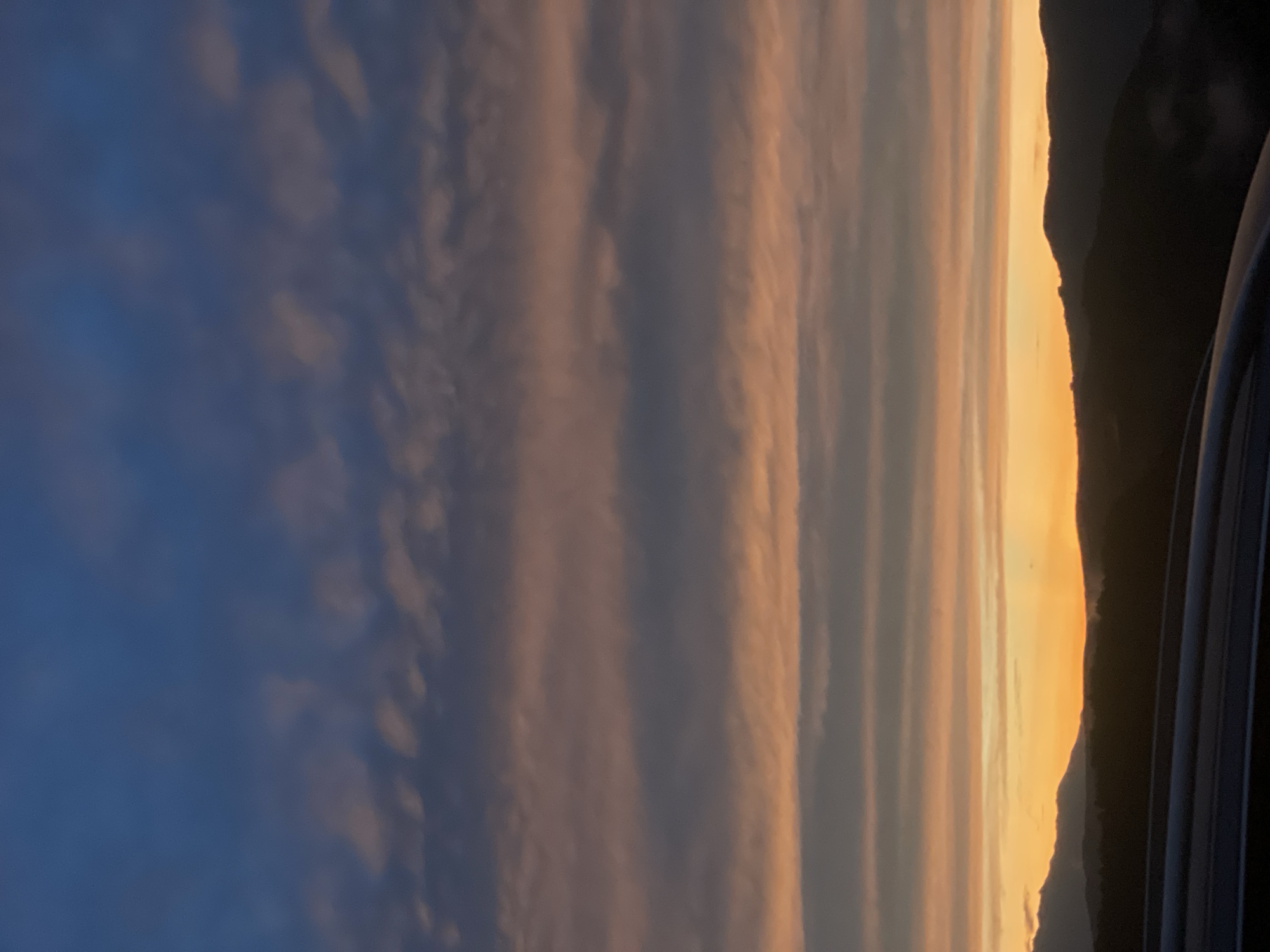 The image depicts a sunset over a hilly landscape, possibly near Bogolin, Bulgaria. The sky is filled with layered clouds in hues of deep blue, gray, and orange, indicating the transition from day to night. The lower part of the image is dominated by silhouetted hills and mountains, with dark trees and vegetation visible against the vibrant sky. In the foreground, the dark, curved surface of what appears to be a vehicle's roof or hood is present. No people or specific objects that would identify a precise urban location are visible.