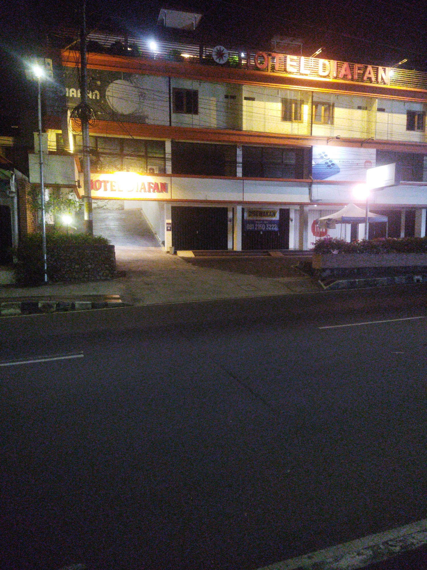 This is a nighttime photograph of the exterior of Hotel Diafan, located on Jl. Jend. Ahmad Yani in the city of Wonogiri, Indonesia. The hotel has a prominent illuminated sign that reads "HOTEL DIAFAN" in bright orange lettering. The building appears to be a multi-story structure with a light-colored facade. In the foreground, there is a dark asphalt road with a white line marking. A sidewalk runs alongside the road, separating it from the hotel. There are streetlights visible, casting a yellow glow on the scene. A stone wall is present near the entrance ramp of the hotel. The overall impression is that of an urban environment at night, with the hotel being the primary subject.