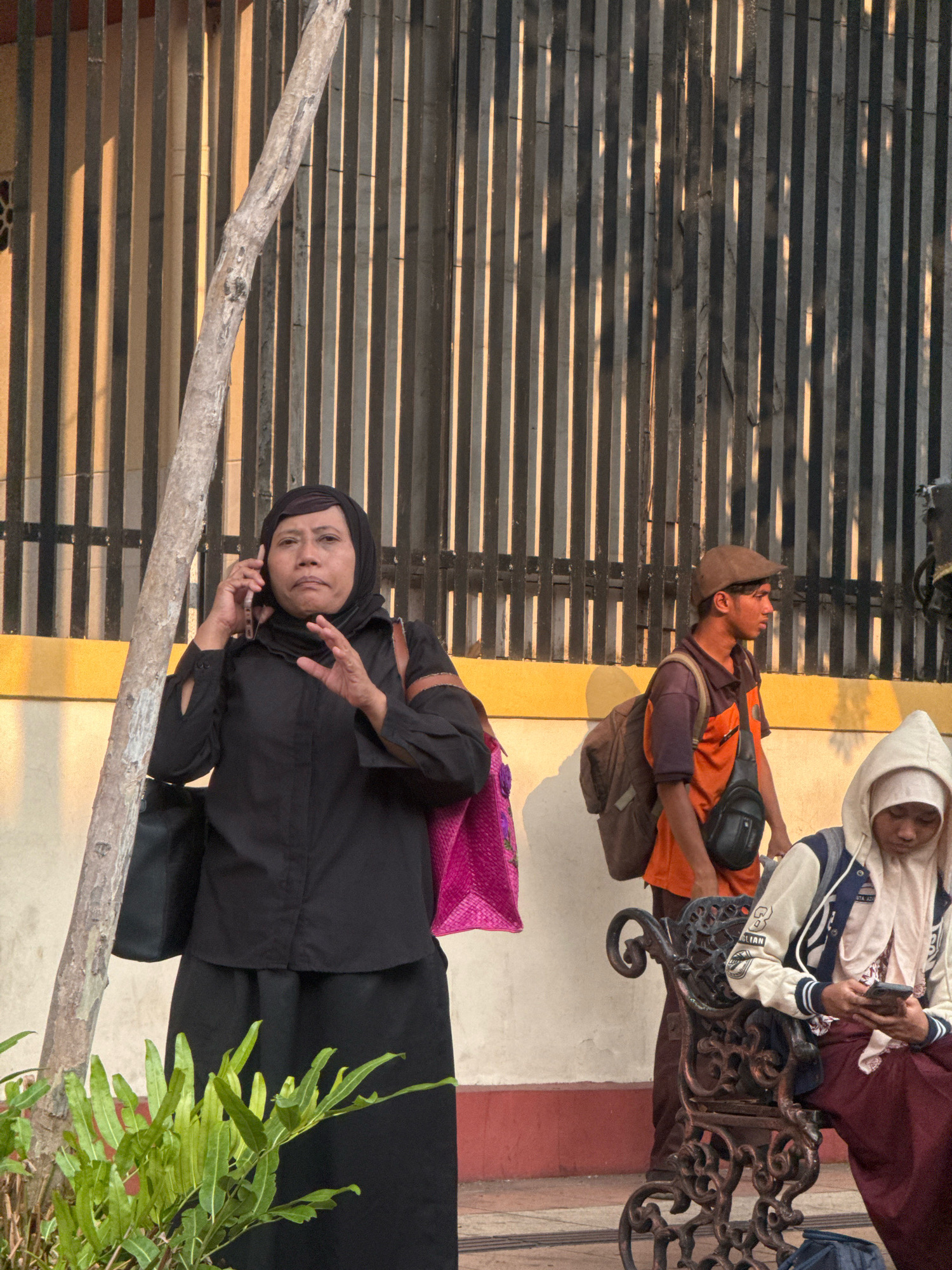 In Surabaya, Indonesia, a woman in a black hijab and dress speaks on a mobile phone, holding a black bag and a pink tote bag. A young man wearing an orange shirt and a brown cap with a backpack walks past. To the right, a person in a white hoodie sits on a dark metal bench, looking at their phone. A tree trunk is visible on the left, and a vertical slatted fence forms the background. The scene appears to be outdoors, possibly near a public area or transportation hub, with warm sunlight casting shadows.