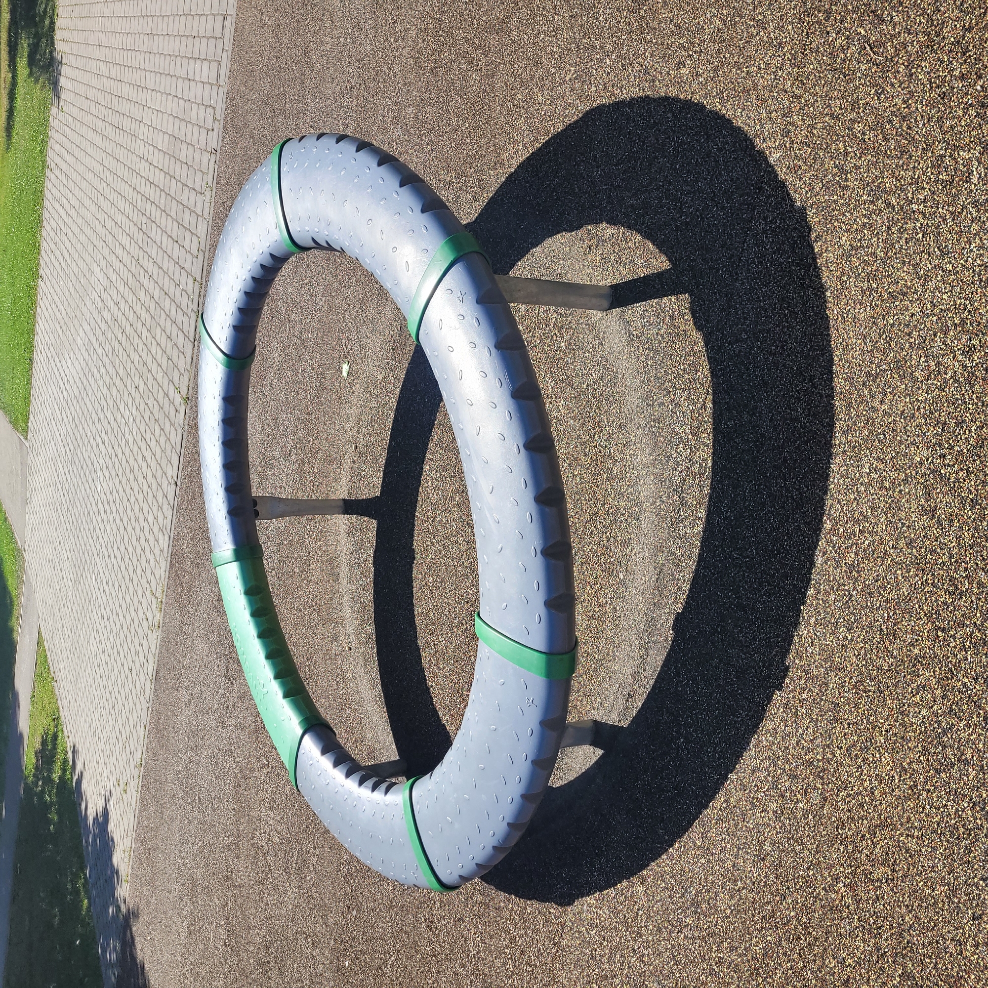 An oval-shaped playground merry-go-round, predominantly grey with green accents, rests on a rubberized surface with a gravel-like texture. The merry-go-round is supported by several metal legs that cast long shadows on the ground. In the background, a paved walkway made of interlocking grey bricks leads to a patch of green grass. The scene is set in an outdoor urban environment, possibly a park or playground. The image does not contain any people or any discernable action. The specific location in Jūrmala, Latvia, cannot be identified from the visual information alone.