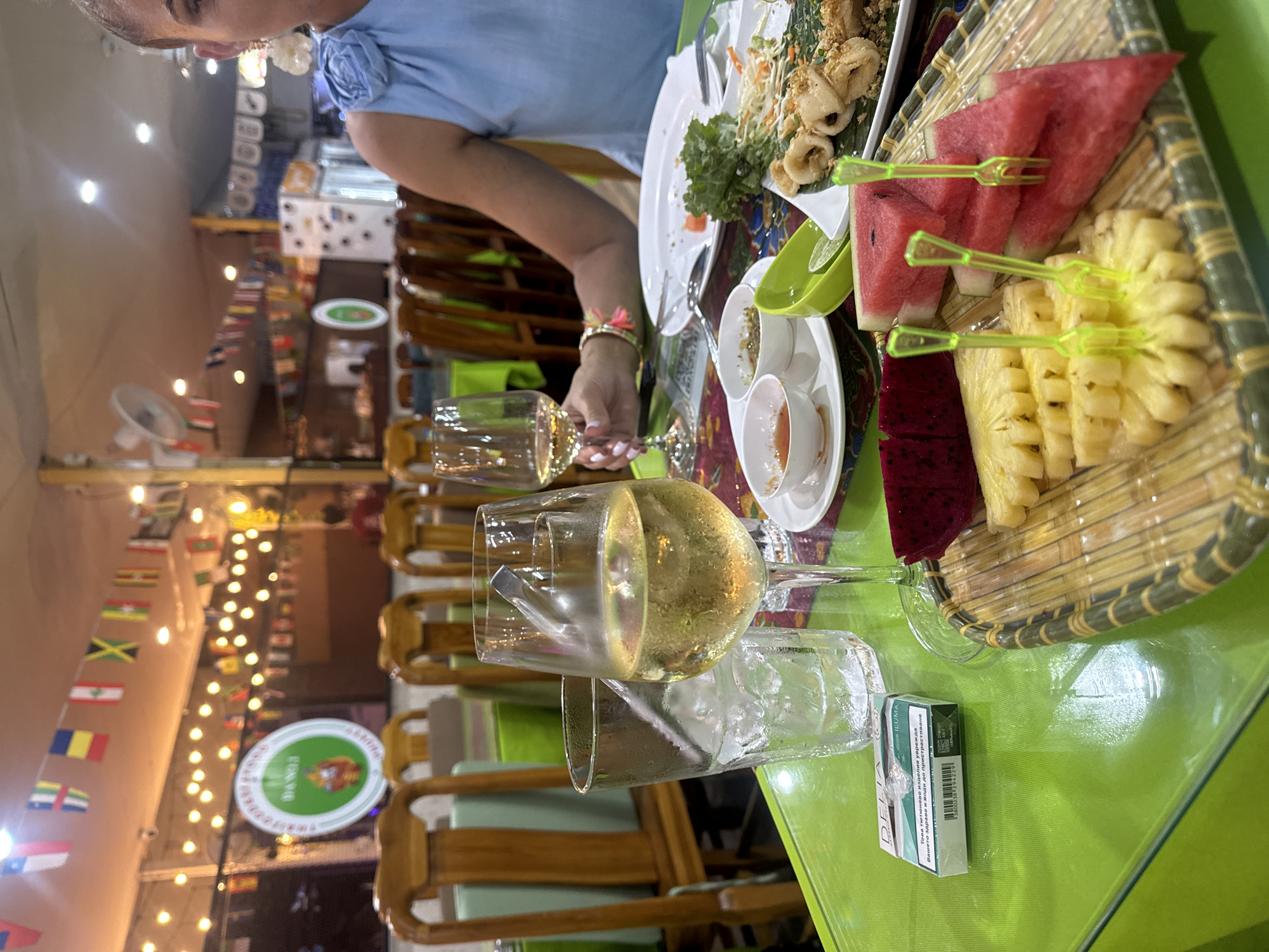 The image captures a scene at a restaurant in Patong, Thailand. A person wearing a light blue shirt is seated at a table with an assortment of food and drinks. The food includes a platter of mixed fruits (watermelon, pineapple, dragon fruit) and another dish that appears to be fried calamari with salad. There are also small dishes containing sauces or dips. A wine glass filled with a light-colored liquid, presumably wine, is prominently displayed on the table, along with an empty glass containing ice. A pack of Delta ILUMA cigarettes is also on the table. The restaurant's interior is decorated with strings of lights and flags from various countries. Several empty wooden chairs are arranged around the table.