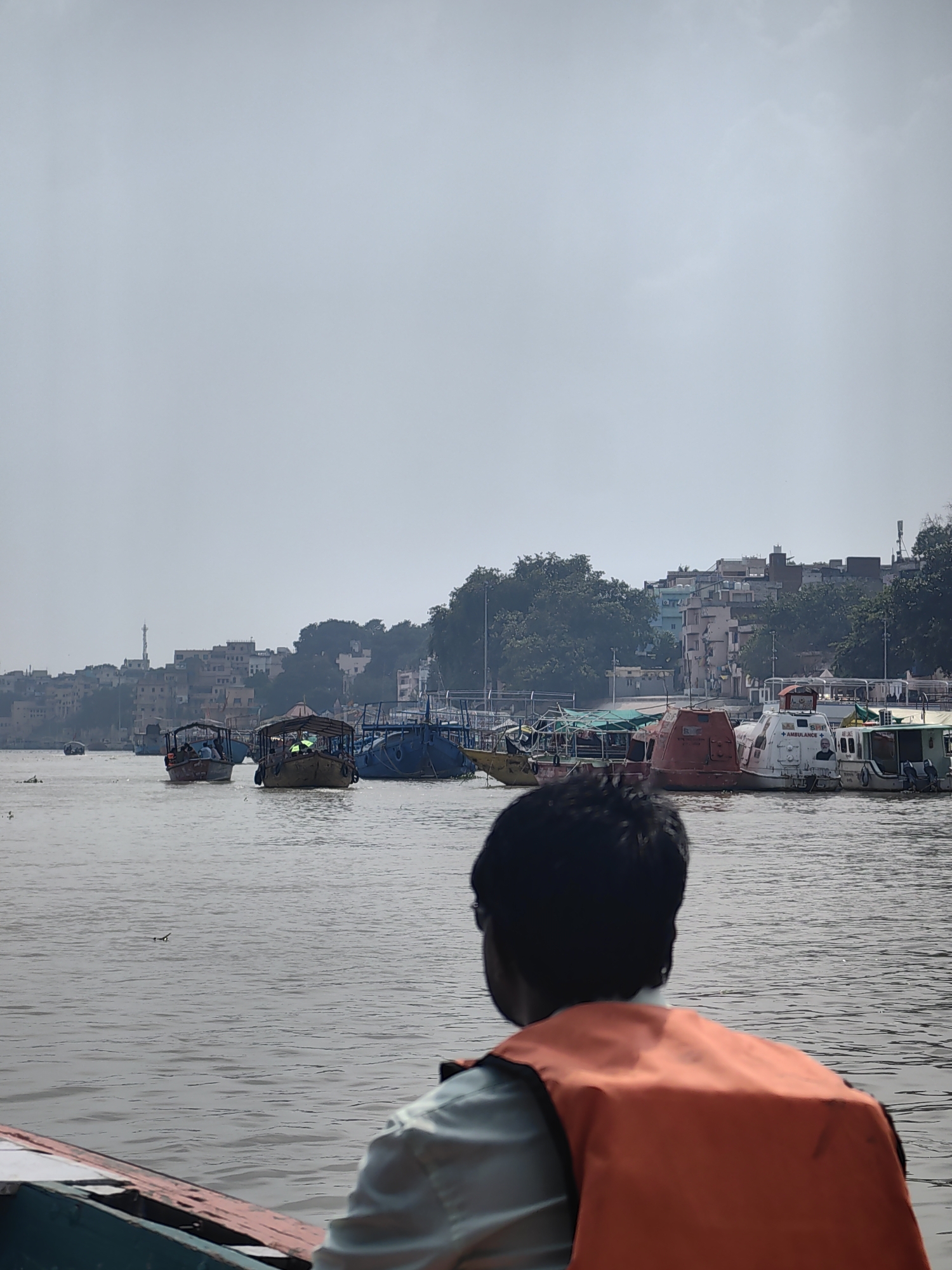The image shows a person wearing an orange life jacket in the foreground, seen from behind, looking out at a river. Several boats are visible on the water. In the background, a city skyline with buildings and trees lines the shore. The sky is overcast. The scene appears to be a view from a boat on the Ganges River in Mughalsarai, India.
