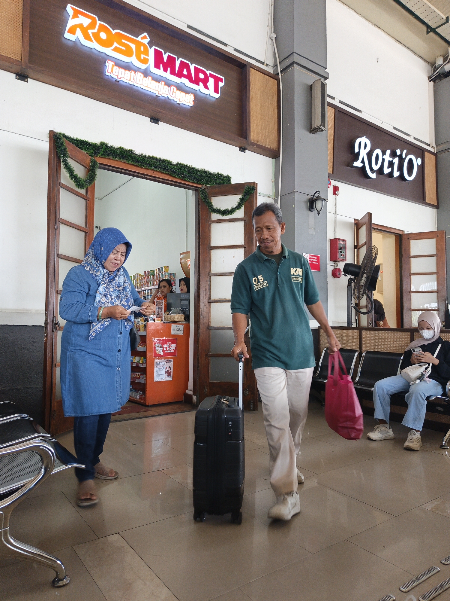 The image depicts the interior of a train station in Klaten, Indonesia. In the foreground, a man in a green polo shirt with "KAI PORSE" and "05 RUBIYO" printed on it, is walking while pulling a black suitcase and carrying a large red tote bag. He is wearing light-colored pants and white sneakers. To his left, a woman wearing a blue denim dress and a blue headscarf is standing in the doorway of a store labeled "ROSE MART" and "Tepat Belanja Cepat". She is holding a small white item, possibly a receipt or ticket. Inside the store, there are shelves stocked with products and two individuals are visible behind a counter. Further back, another woman is seated on a bench, looking at her phone. The environment appears to be a waiting area with tiled flooring and wooden doors and trim. Above the "ROSE MART" store, a neon sign displays the store's name, and to the right, another neon sign reads "Roti'o". Green tinsel is draped around the entrance of the "ROSE MART". A black oscillating fan is mounted on a post.