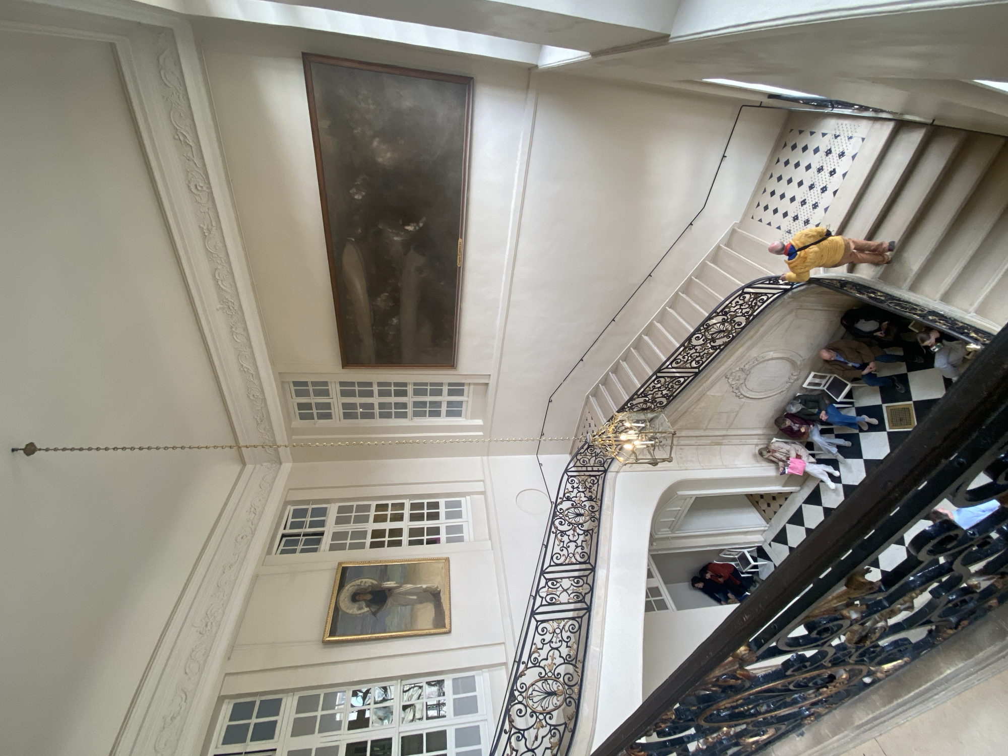 The image depicts a grand interior staircase within the Musée d'Orsay in Paris, France. The multi-level structure features off-white walls, light-colored steps, and ornate black wrought iron railings with gold scrollwork. Numerous tall, rectangular windows with white frames are visible on the left and right walls, admitting natural light. A multi-bulb, caged chandelier hangs from a long chain in the center of the stairwell.
On the upper left wall, a vertical framed painting, identifiable as "Femme à l'ombrelle" by Henri-Edmond Cross, portrays a woman on a beach. On the upper right wall, a large, horizontal framed painting with a dark, muted palette depicts a landscape or historical scene, with a small label visible beneath it.
The floor sections between the stair flights feature a black and white checkered tile pattern. Several individuals are present: five are seated on white chairs on a lower landing, with two in a recessed alcove and three closer to the main staircase. Another person is partially visible seated on a lower step in the foreground. On the main staircase, a person wearing a bright yellow jacket, blue backpack, and a multi-colored beanie stands facing away from the viewer, gesturing upwards with their right hand.