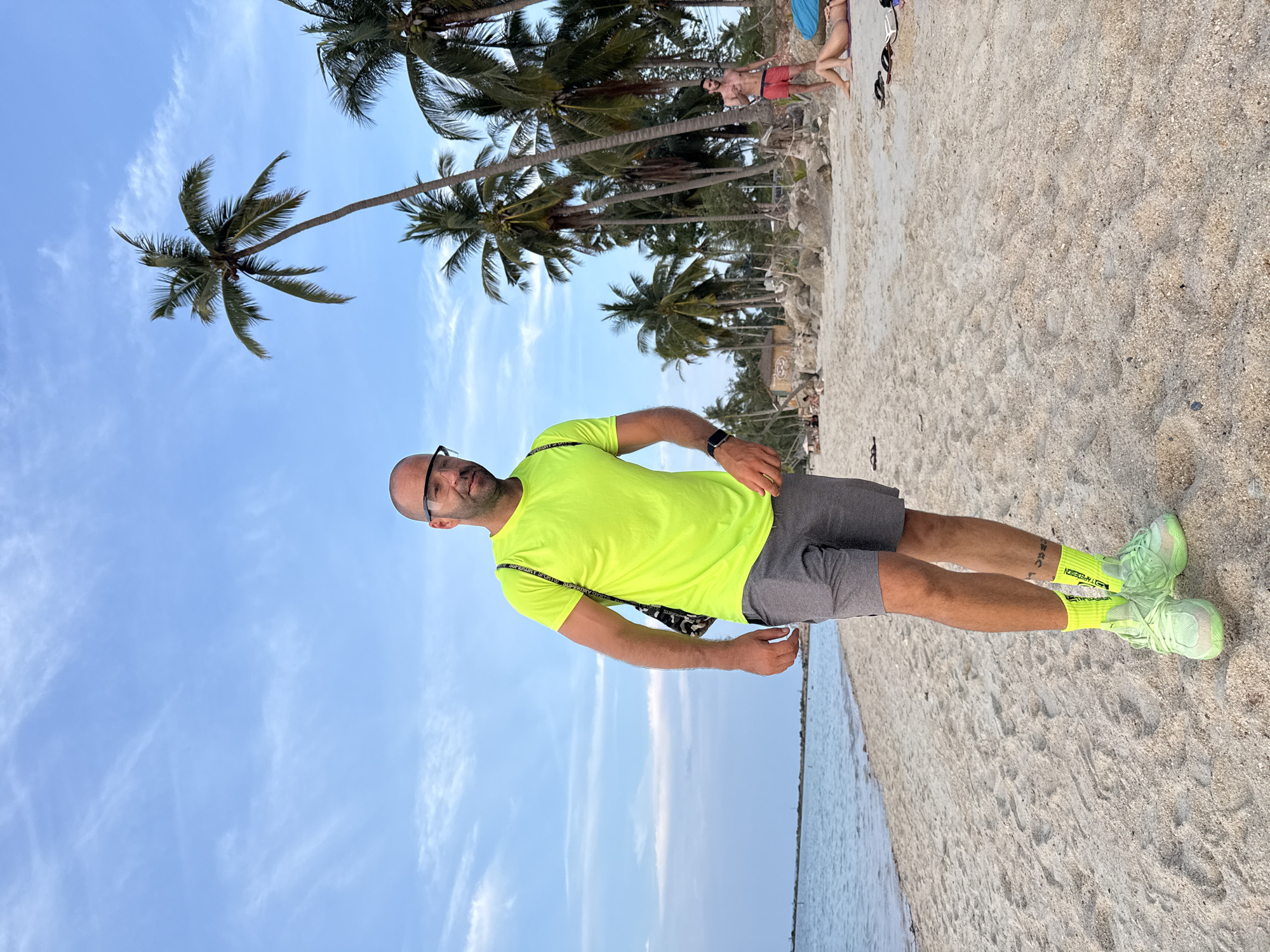 A man stands on a sandy beach in Ko Samui District, Thailand, wearing a bright neon yellow t-shirt, gray shorts, neon green socks, and matching neon green sneakers. He has a bald head and is wearing clear glasses. A patterned bag is slung over his left shoulder. The beach has scattered palm trees and a calm ocean in the background. Other people are visible further down the beach, some sitting and others walking. The sky is blue with scattered white clouds.