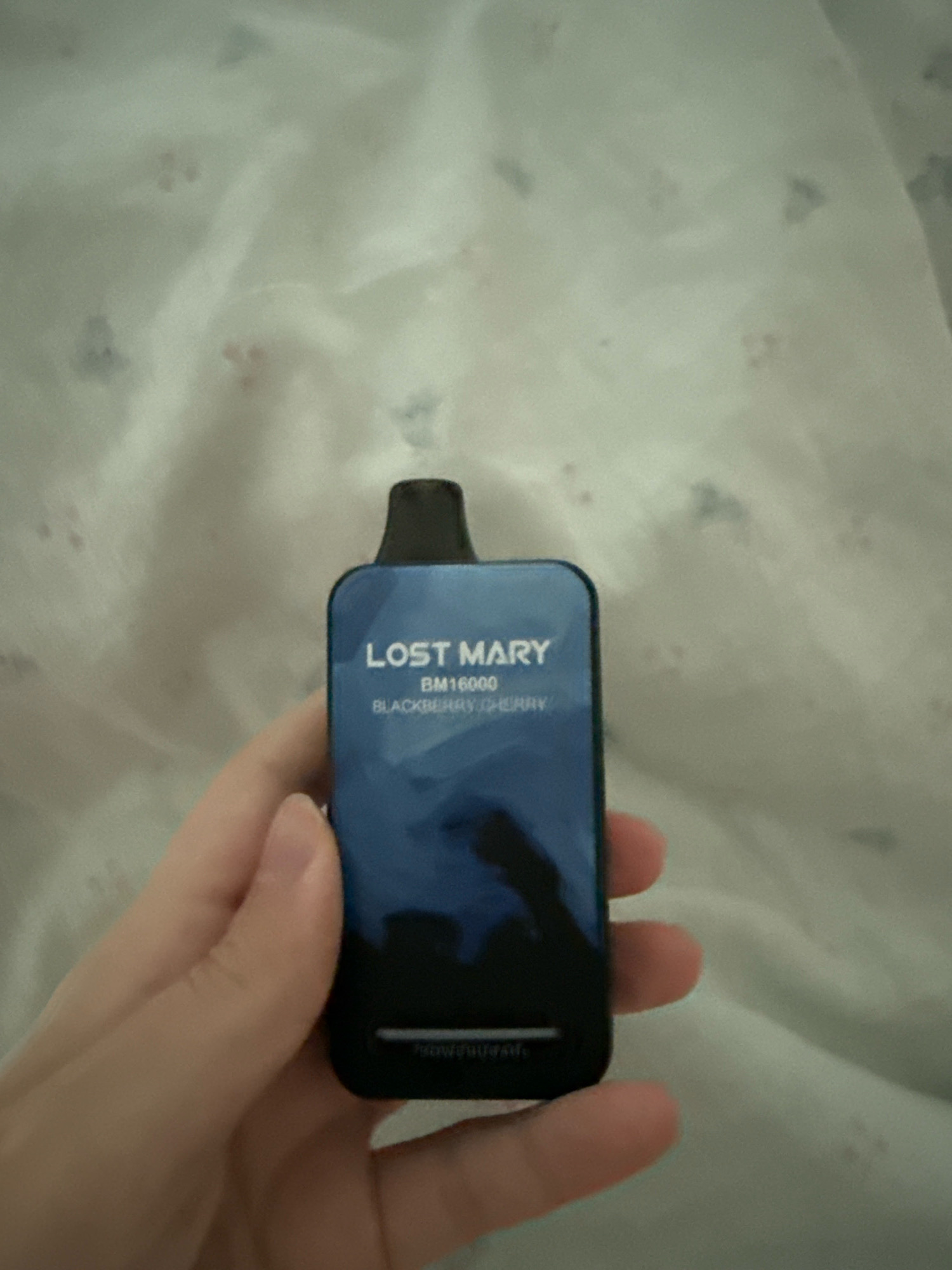 A hand holds a blue disposable electronic cigarette in front of a white, patterned fabric background. The electronic cigarette has "LOST MARY" printed in white text at the top, followed by "BM16000" and "BLACKBERRY CHERRY". The fabric appears to be a duvet or blanket with a repeating pattern of small, dark shapes that resemble paw prints. There is no indication of location in Zavolzhye, Russia or any other city.