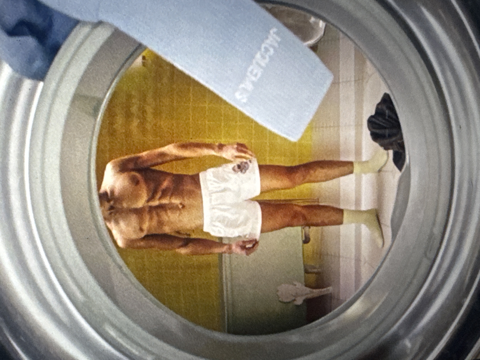 The image shows a man inside what appears to be a washing machine drum, viewed from the outside. He is wearing white boxer shorts and light-colored socks. His body is positioned horizontally, and he is looking towards the viewer. His arms are bent, and his hands are near his waist. A blue fabric with the word "JACQUEMUS" in white letters is draped over the top edge of the washing machine drum, partially obscuring the man's head. The background within the drum appears to be a yellow tiled wall, and to the right, a black object resembling a laundry bag or clothing is visible. The overall scene is indoors, possibly a laundry room or bathroom, given the presence of the washing machine. There is no indication of time of day or weather. The man's facial expression is not clearly discernible. The location context of Kuta Selatan, Indonesia, does not provide any direct visual cues in the image itself.