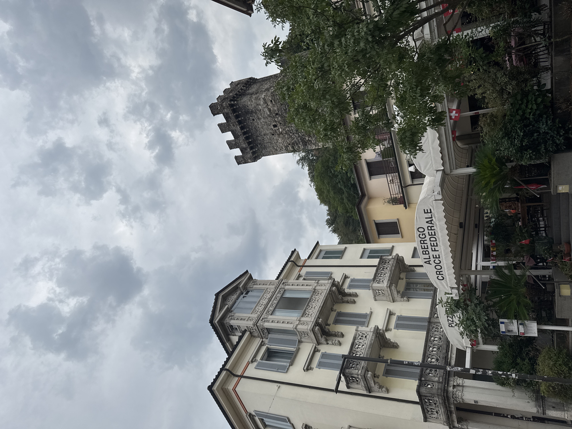 The image shows a view of the Castel Grande in Bellinzona, Switzerland, under a cloudy sky. To the left, a light-colored building with multiple balconies and gray shutters is visible. The ground floor of this building features a white awning with the words "ALBERGO CROCE FEDERALE" and "PIZZERIA" printed on it, suggesting it is a hotel and pizzeria. Below the awning, there are tables and chairs for outdoor dining, along with lush green plants. To the right of this building, the stone tower of Castel Grande rises above the trees. The tower has a crenellated top and appears to be made of dark stone blocks. Further to the right, another building with yellow walls and dark windows is partially visible. Several small Swiss flags are displayed on poles and on the awnings of the establishment.