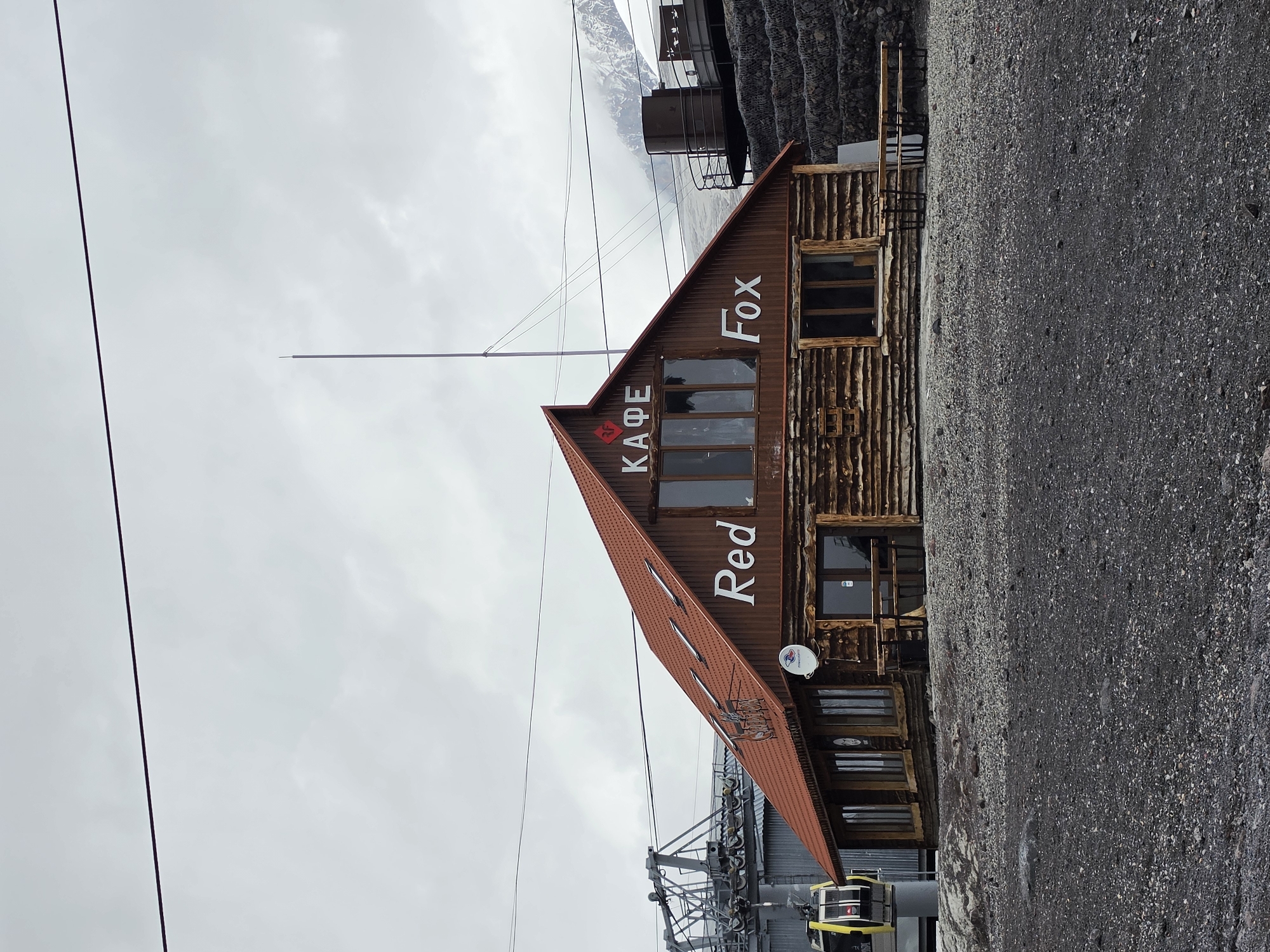 A rustic wooden cafe, labeled "КАФЕ Red Fox," stands on a gravelly, rocky terrain. The cafe has a steep, red-tiled roof with several skylights and a prominent "RF" logo above the entrance. Large windows reveal the interior, and a satellite dish is mounted on the exterior wall. In the background, a ski lift structure with yellow gondolas is visible, suggesting a mountain resort setting. The sky is overcast, and distant snow-capped mountains can be seen. The cafe is empty of people, and no activity is taking place. The scene appears to be in the city of Terskol, Russia, a popular destination for mountain sports and tourism.