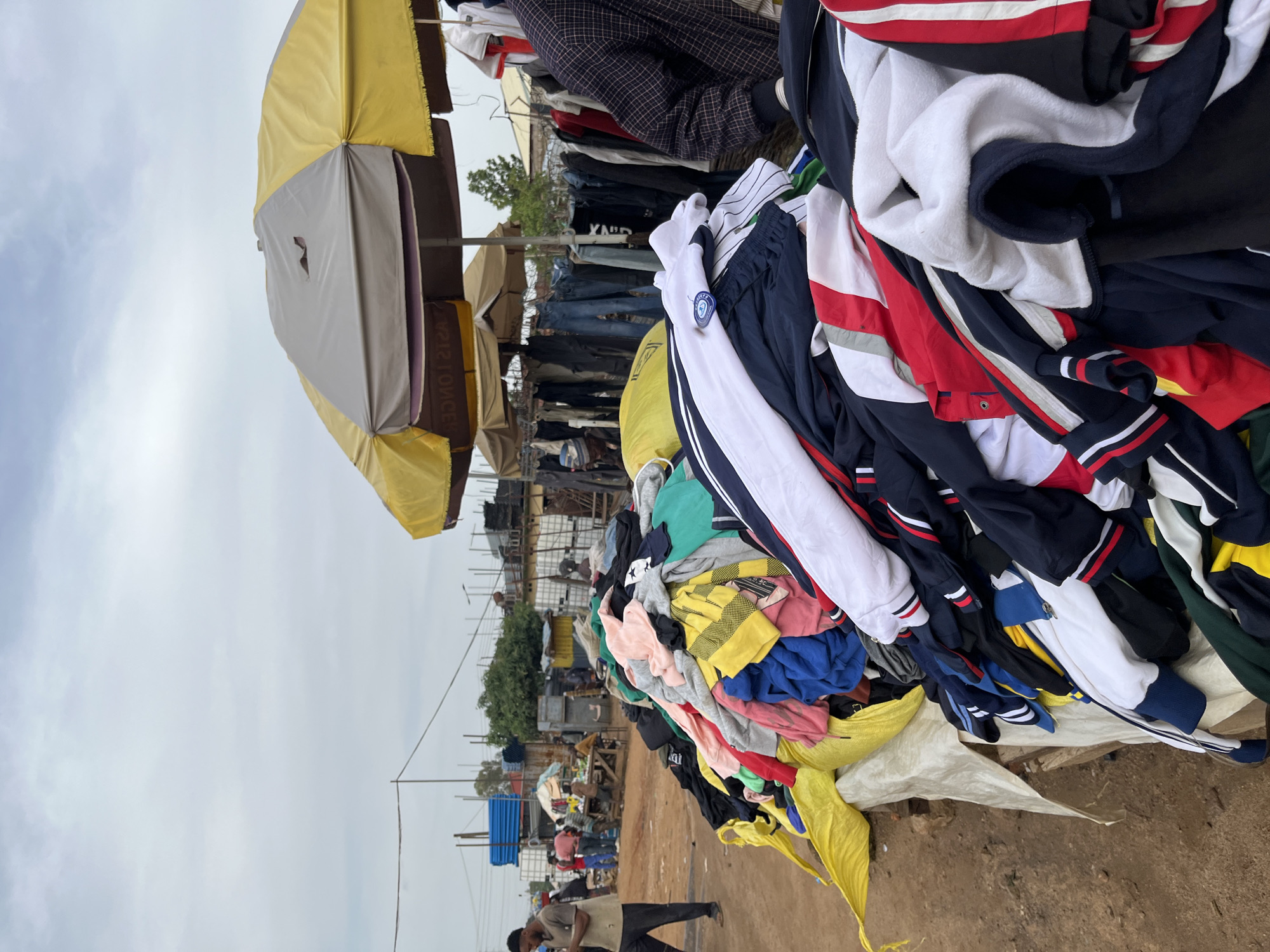The image depicts an outdoor market scene in Gura Top, Nigeria, with a focus on a stall selling clothing. The primary subjects are piles of colorful clothing, predominantly jackets and pants, neatly folded or draped. Several umbrellas, one large yellow and grey umbrella being prominent, provide shade over the market stalls. In the background, there are glimpses of other market stalls, metal structures, and a few individuals, suggesting a bustling commercial environment.
The activity taking place is the selling and likely browsing of second-hand clothing. The items are displayed in a somewhat organized yet abundant manner, indicative of a typical outdoor clothing market. The time of day appears to be daytime, with overcast skies contributing to a diffused light. The weather seems to be warm and potentially humid, common for an outdoor market setting in Nigeria.
Notable details include the variety of clothing styles and colors, with some pieces featuring stripes and prominent brand logos or emblems, such as a blue badge on a white and navy garment. The ground is unpaved, typical of many market areas in developing regions. The general atmosphere conveyed is one of everyday commerce and resourcefulness, with people engaging in economic activities. There is no visible text in the image that is clearly legible.