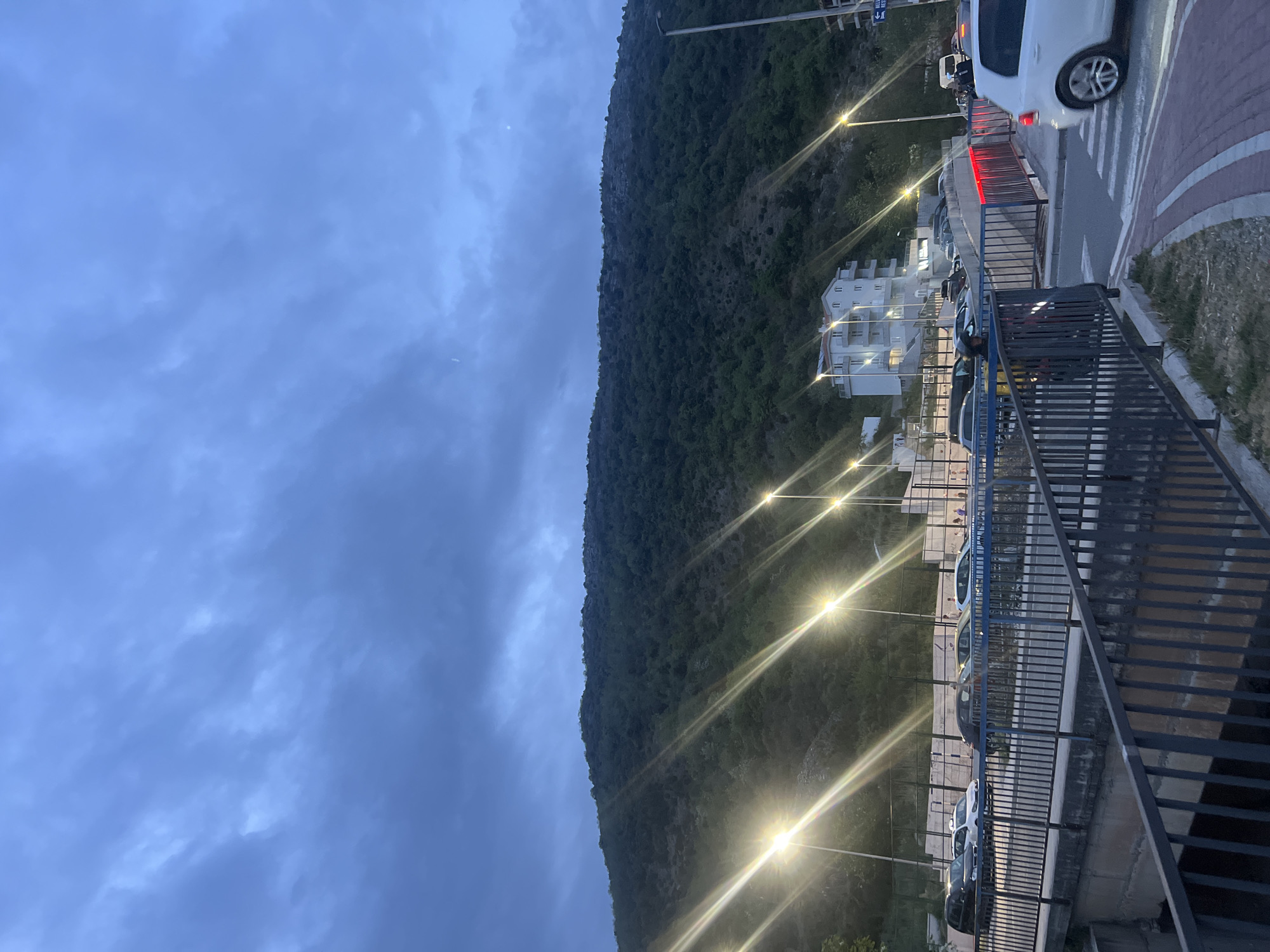 The image depicts a view in Boreti, Montenegro at dusk. A large, green mountain dominates the view, rising sharply from the right side of the frame. The sky is overcast with dark blue and gray clouds. Streetlights line the road that runs along the base of the mountain, casting a bright glow. Parked cars are visible along the road. A white building with multiple stories is situated near the middle of the frame. A pedestrian walkway with metal railings is located on the right side of the image. A white car is partially visible on the right, near a crosswalk with red-and-white brick patterns. The scene appears to be generally deserted of people.