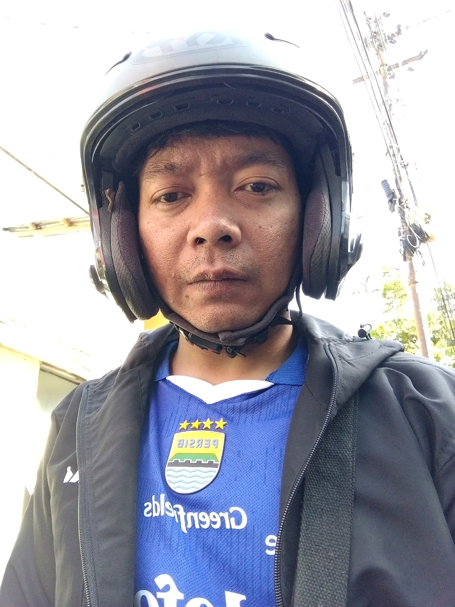 An adult male wearing a black helmet and a blue shirt with a crest and text on it, is pictured. He is also wearing a black jacket. The image appears to be taken outdoors, with a blurred background of trees and power lines, suggesting an urban or suburban setting. The lighting is bright, and the image has a slight overexposure. The man's expression is neutral. The crest on the shirt features waves and stars, and some text which is partially reversed due to the image being a selfie. The overall scene is casual and informal, possibly depicting someone preparing for or returning from a motorcycle ride. The provided text "Persib" on the shirt is a strong indicator that this photo may have been taken in Bandung City, Indonesia, as Persib is a very popular football club from that region.