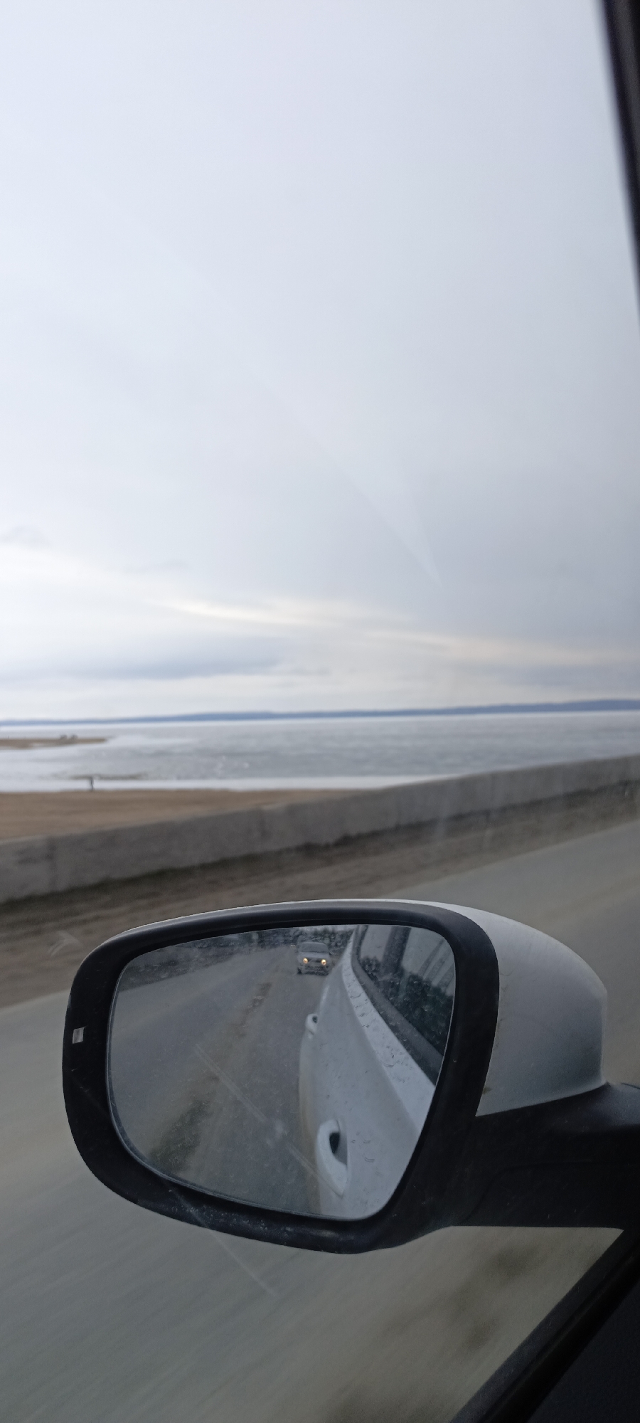 Captured from within a moving vehicle, the photograph depicts a landscape viewable through the car's side mirror. In the foreground, the side mirror reflects the adjacent lane, showing a white car and another vehicle further behind with its headlights visible. The primary focus is on the wider external scene: a vast expanse of water or partially frozen lake extends to the horizon, merging with a clouded sky. A low concrete barrier or embankment runs along the edge of the waterway. The overall light suggests an overcast day. This could possibly be located on a body of water in Russia.