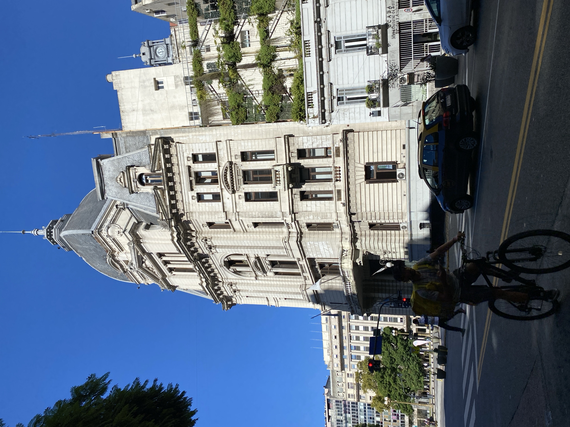 This image depicts a sunny street scene in Buenos Aires, Argentina, showcasing a blend of architectural styles under a clear blue sky.
In the foreground and prominent on the left is a magnificent, ornate building constructed of light-colored stone, featuring classical architecture with multiple stories, decorative cornices, arched windows, and a distinctive domed roof topped with a spire. Directly behind it, another building rises, characterized by modern design elements including a section covered in lush green vertical gardens arranged on trellises. A clock tower with a dome is visible above this structure.
On the right side of the street, another light-colored building has balconies and shutters. Visible text on this building identifies it as "GCABA" (Government of the City of Buenos Aires) and "Mesa General de Entradas y Archivo" (General Entry Desk and Archive), confirming the location.
The street itself is asphalt with clear double yellow lines and crosswalk markings. A dark-colored taxi with yellow accents, characteristic of Buenos Aires, is visible on the road. In the foreground, a person wearing a helmet and a light-colored top is riding a bicycle towards the right side of the frame. Further down the street, more urban buildings, green trees, and a red traffic light are visible.
The scene is bathed in bright sunlight, indicating a clear day, likely mid-morning or early afternoon, with distinct shadows being cast. The overall impression is that of a vibrant and active city environment.