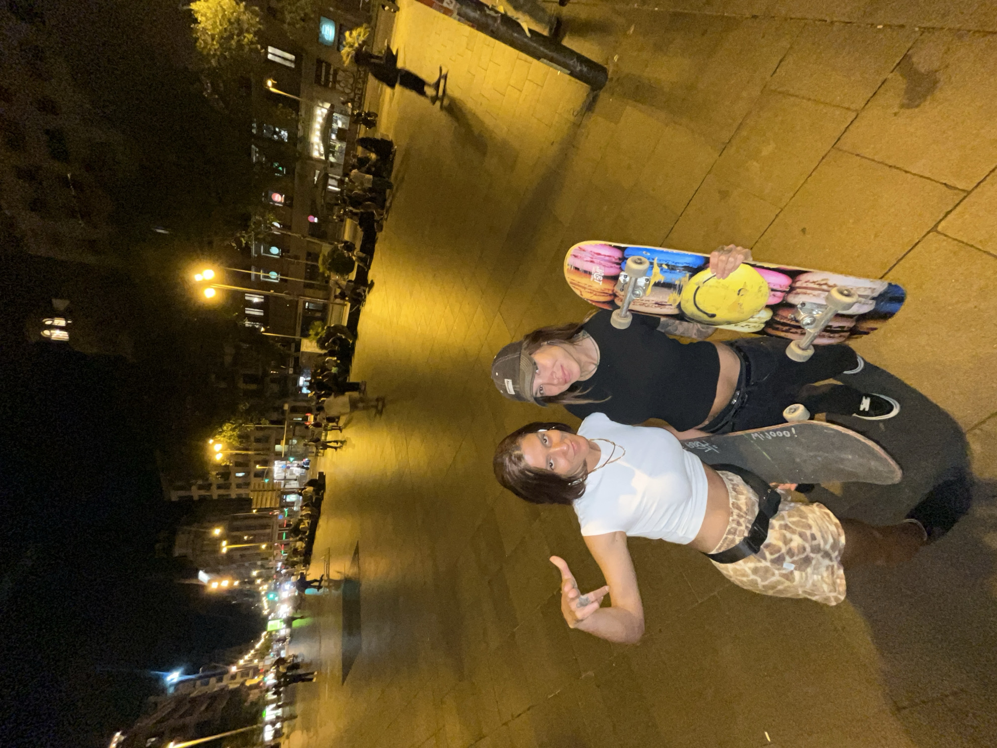 This is a nighttime, high-angle, full-shot photograph taken in Barcelona, Spain, on Las Ramblas. Two women are in the foreground, holding skateboards. The woman on the left wears a white crop top, leopard-print shorts, and black socks. She is making a hand gesture. The woman on the right wears a black t-shirt, black pants, and a baseball cap. She is holding a colorful skateboard adorned with a smiley face and macarons. The background shows a street scene with people walking, sitting, and a few on skateboards. Buildings are visible on either side of the street, lit by streetlights. The ground is paved with stone.