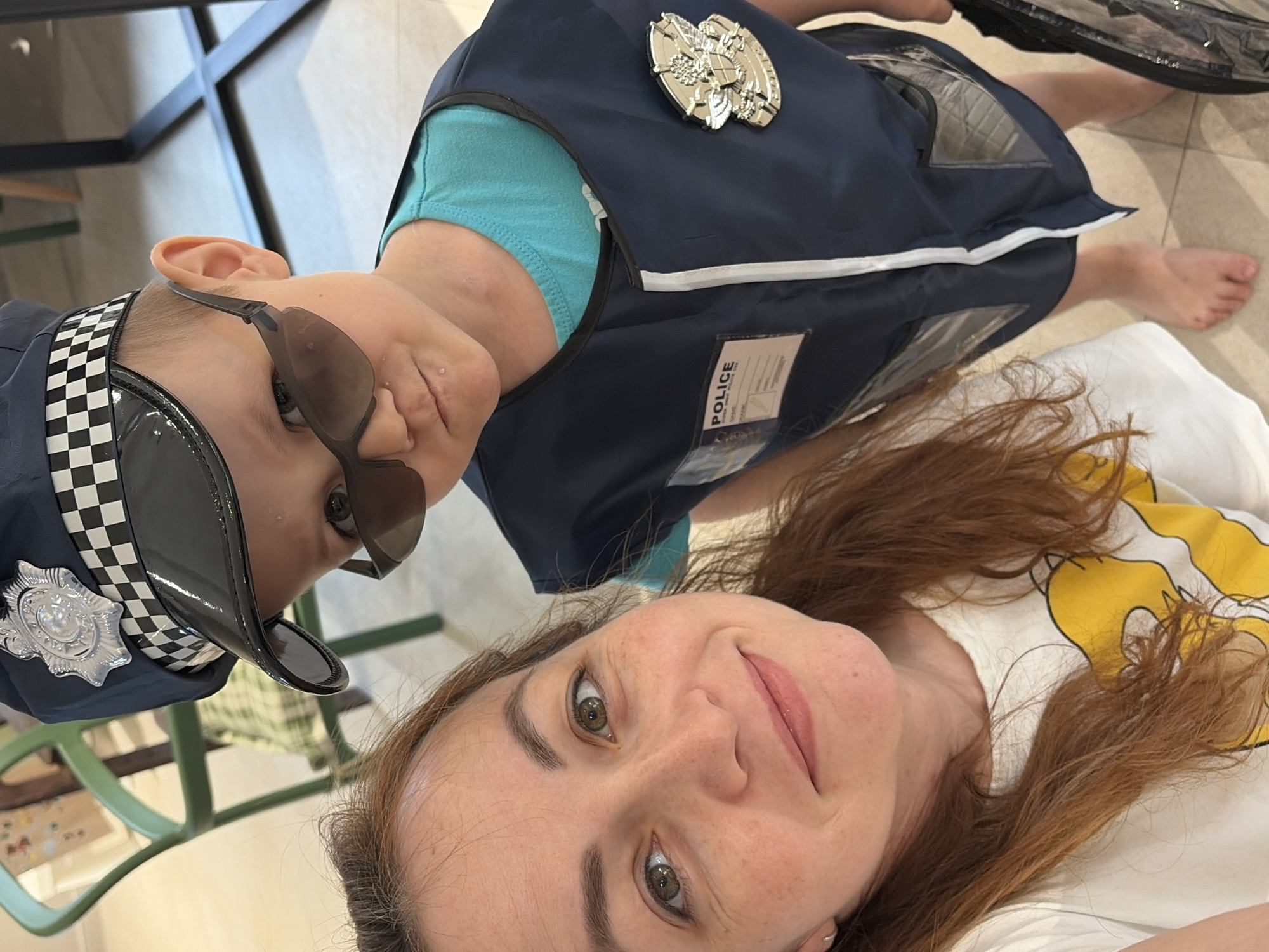 A selfie photograph captures a woman and a child in costume. The child is dressed in a police uniform, complete with a hat, sunglasses, and a police badge on the chest. The woman is wearing a white T-shirt with yellow designs. They are indoors, with furniture visible in the background. The location is not evident from the image and cannot be determined to be Chaikovsky, Russia, without additional information.