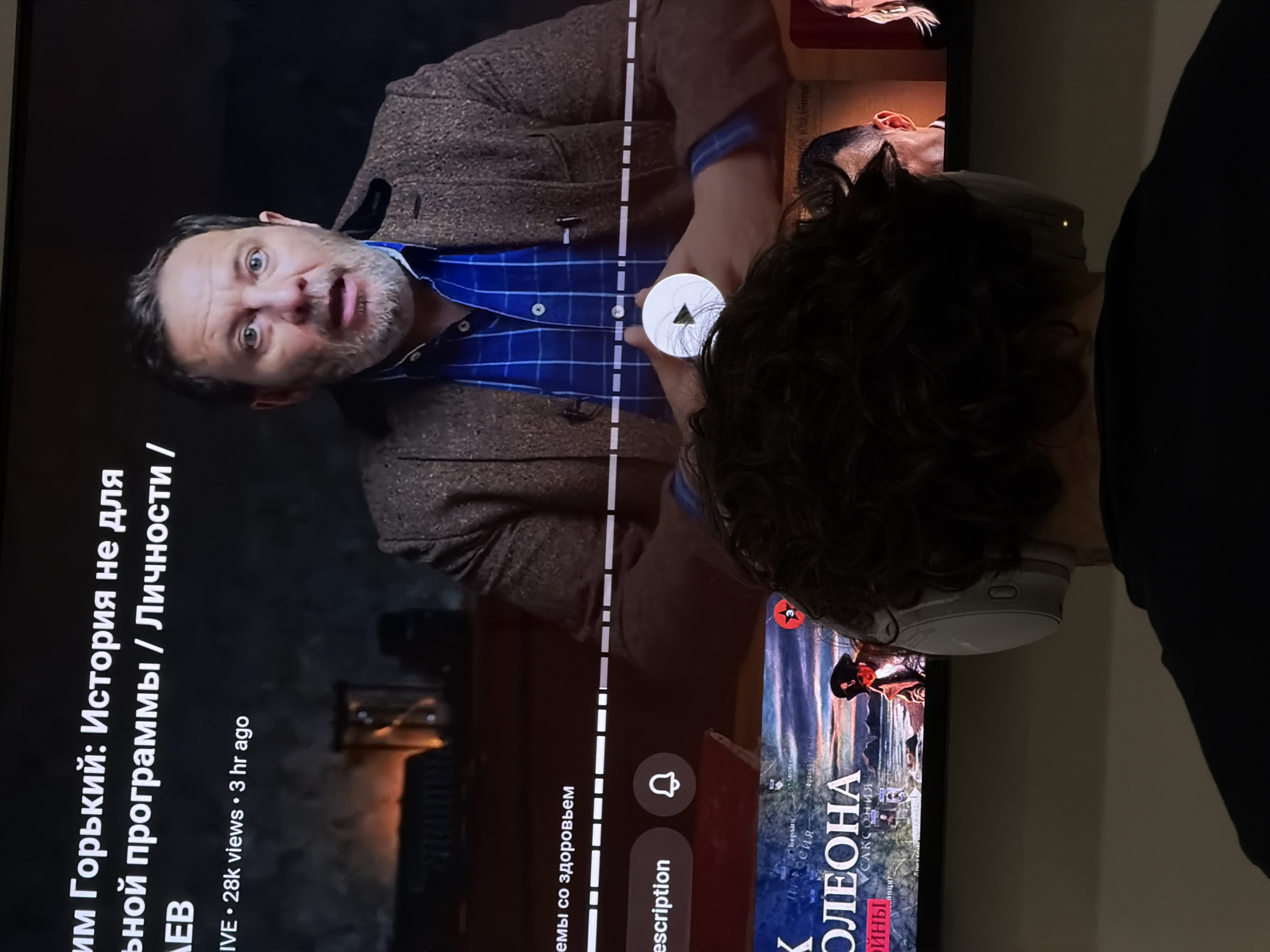 A person with curly hair is watching a video on a TV screen. The video appears to be a documentary or historical program about Maxim Gorky, the famous Russian writer. The person is wearing headphones and is close to the screen. The scene also displays elements of the YouTube interface such as the play button, like and share buttons, as well as the channel name. The image does not contain data that allows for the identification of the location as North Kuta, Indonesia.