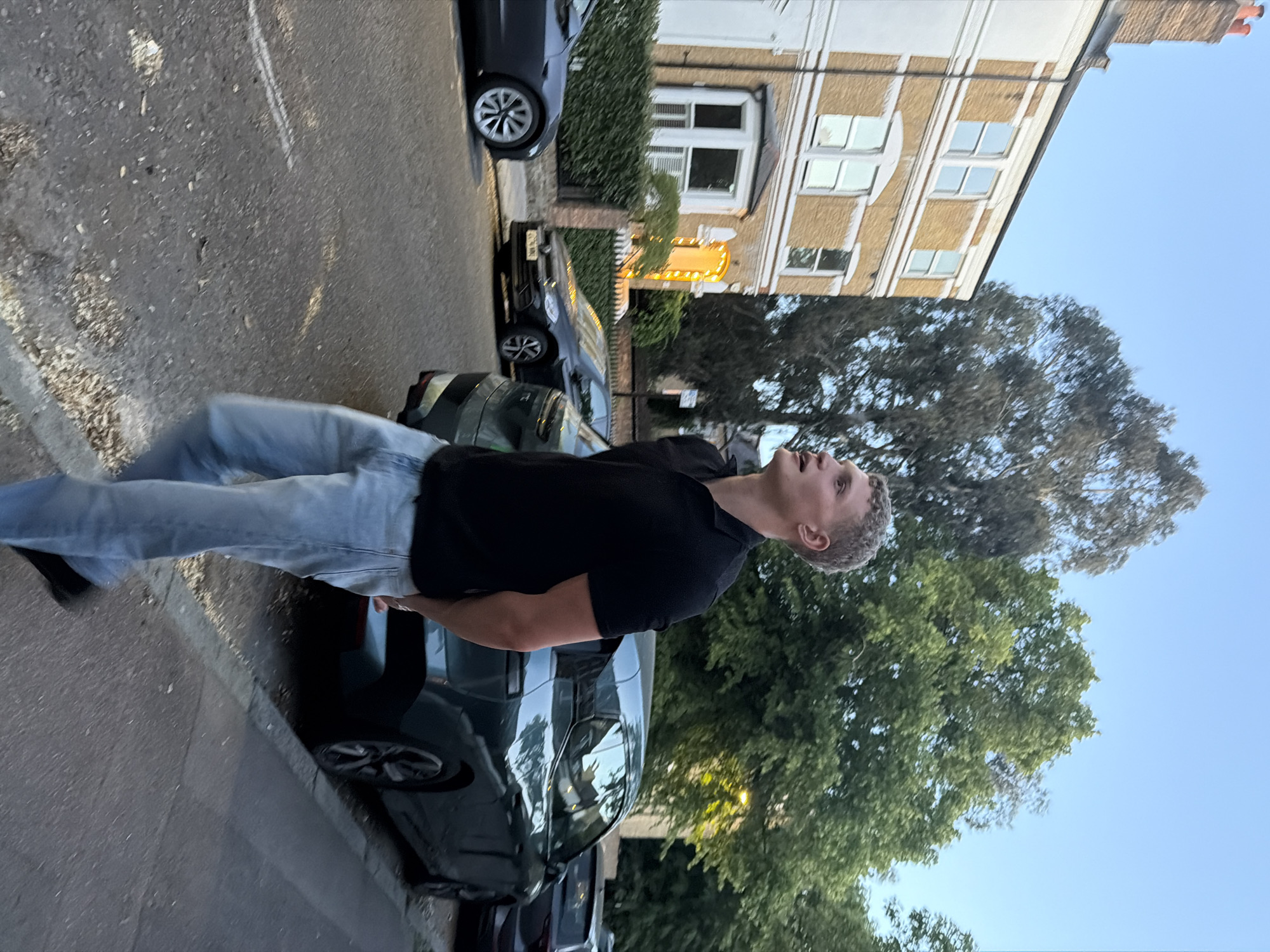 A medium shot depicts a Caucasian male with short blonde hair, clad in a black T-shirt and light-wash jeans, standing outdoors, facing upwards and to the right, seemingly observing something above. The setting appears to be a street in London, United Kingdom, lined with parked cars and residential buildings. The buildings are traditional London brick homes with white window frames. Trees are visible in the background, along with a clear, blue sky. The man is standing on a paved street, near the curb.