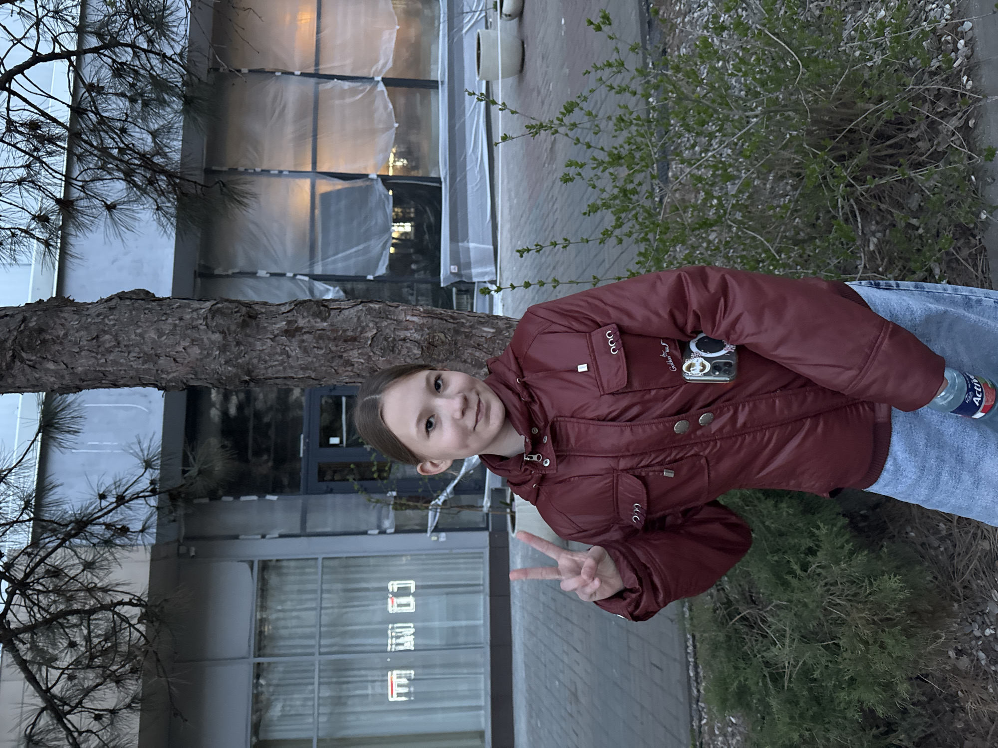 A young person stands in front of a building in what appears to be a city environment. The individual is wearing a reddish-brown jacket and blue jeans, and they are holding up their hand in a peace sign gesture. A water bottle is also visible in their grasp. The building behind them has reflective windows and is partially covered with protective plastic sheeting. A tree is positioned to the left, and some greenery and vegetation are to the right. The location is Azov, Russia.