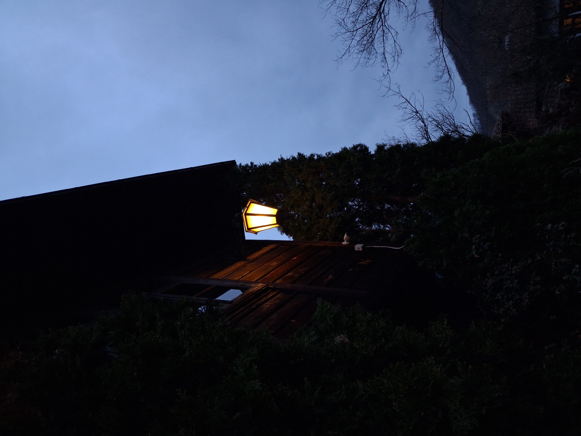 An outdoor scene at night featuring a building with a lit lantern. The lantern is mounted above a window and casts light on the wall beneath it. The structure is partially obscured by surrounding vegetation, and branches from nearby trees are visible against the dark sky. The sky is a mix of dark blue and grey hues. The location is suggested to be Katunishte, Bulgaria.