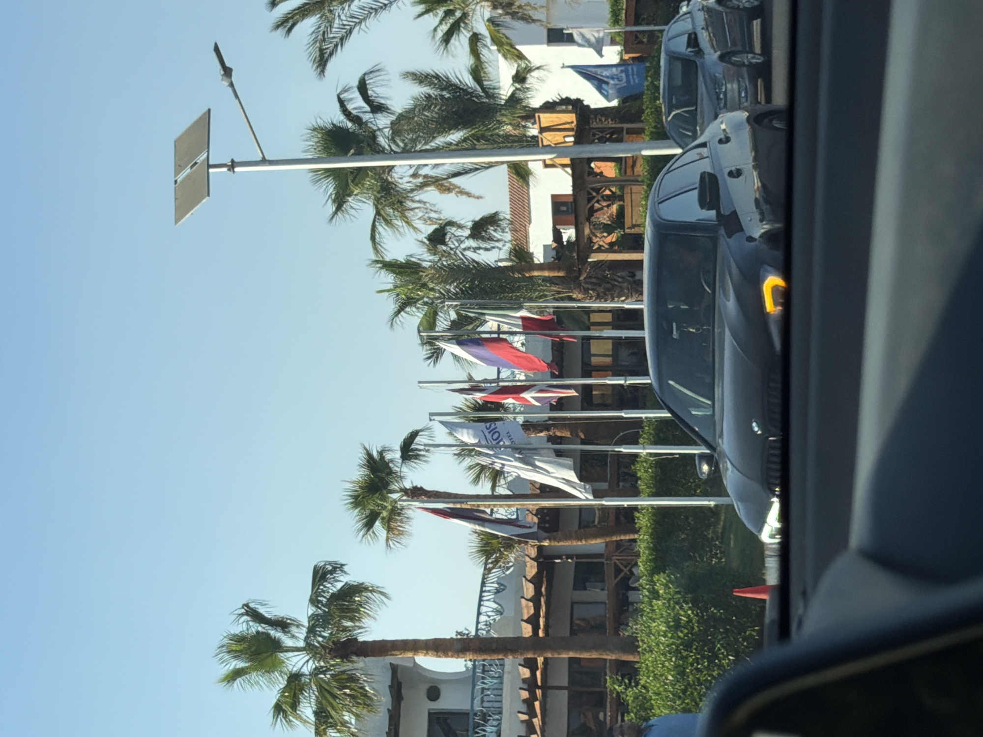 The image shows a street scene in Sharm El Sheikh, Egypt. In the foreground, a dark-colored sedan, likely a BMW, is visible, with its hazard lights illuminated. A gray sedan is behind it. The background features several palm trees, a building with white walls and a red-tiled roof, and an assortment of national flags displayed on poles. A solar-powered street light is also prominent. The sky is clear and blue. The scene appears to be a sunny day, possibly in a resort area given the presence of flags and palm trees.