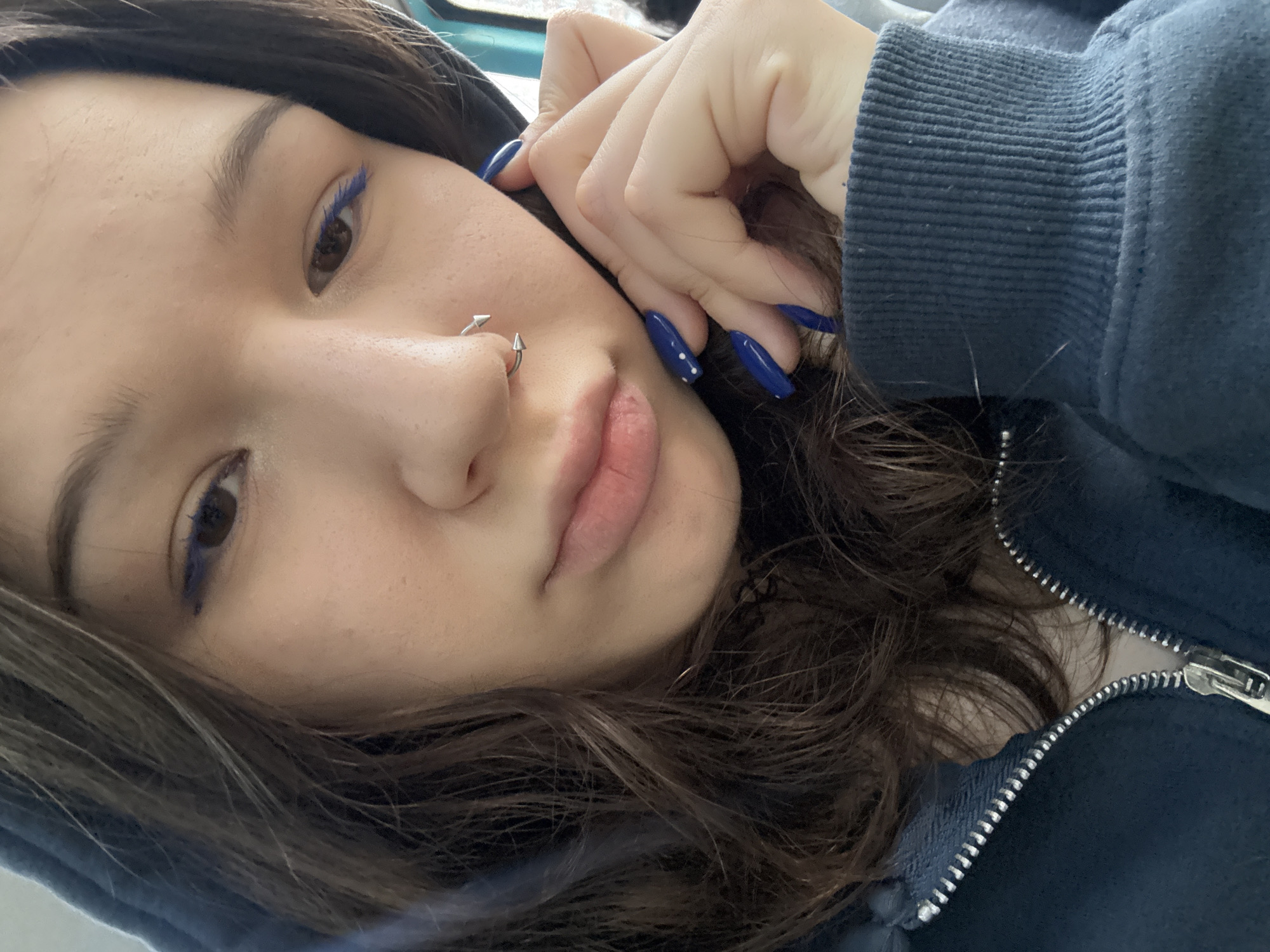 A close-up, eye-level, portrait shot captures a young person from the chest up, looking slightly down and to the right. The subject, who appears to be female, has long, wavy brown hair framing their face. They are wearing a dark blue hooded sweatshirt, zipped partway up. The left side of the subject's face is in focus, revealing dark brown eyes with blue eyeliner on the lower lash line. A silver septum ring with pointed ends adorns their nose. Their mouth is slightly open, and their lips are pink. The right hand is visible, with the fingers resting on the cheek, and the nails are painted a vibrant blue, with white dots on some. The background is blurred, suggesting an indoor setting. The overall mood is introspective and casual. There is no indication of the location within the city of Saint Petersburg, Russia.