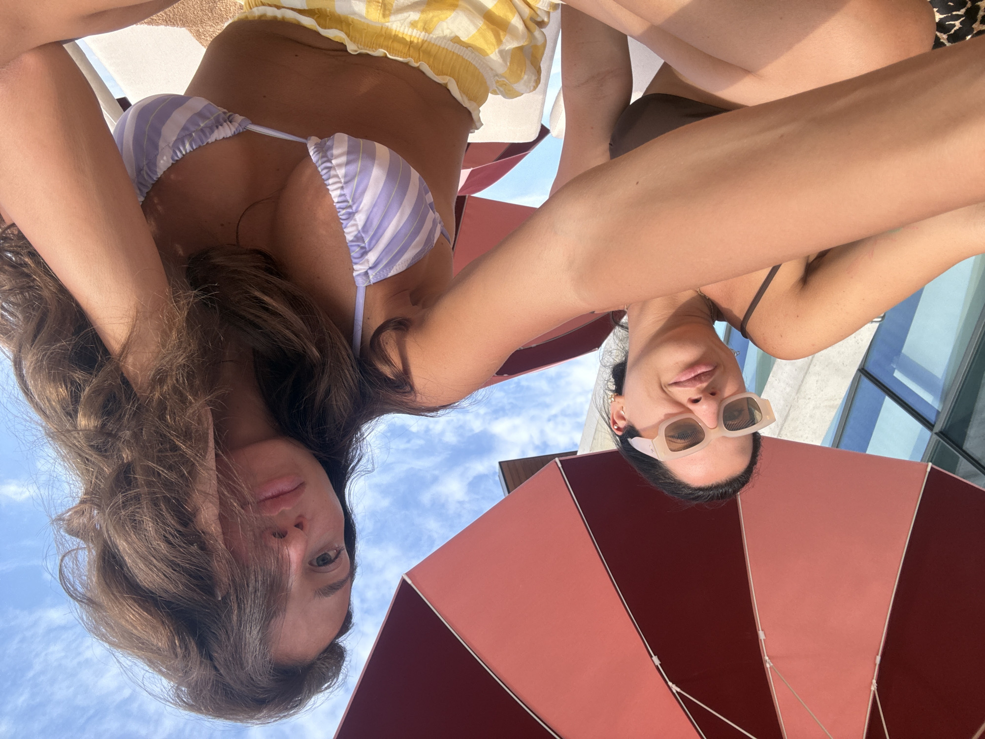 Two individuals are positioned under a large red umbrella. The person on the left wears sunglasses and a brown bikini top, while the person on the right wears a purple and white striped bikini top and yellow and white checkered shorts. The sky is visible behind them, with some clouds. The setting appears to be an outdoor leisure area, possibly a poolside or beach. There is no discernible indication that this scene is located in Sighnaghi, Georgia.