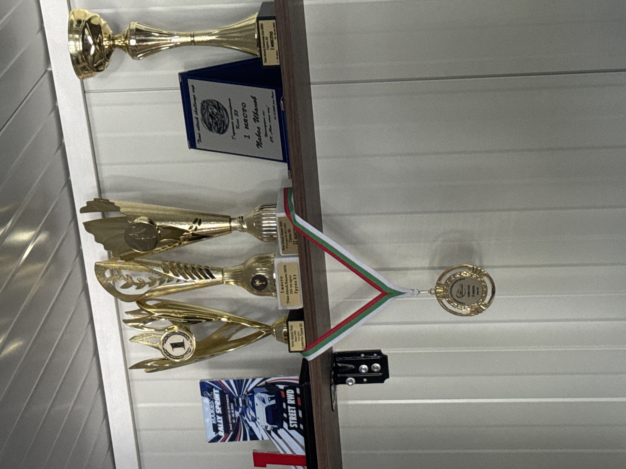 On a wooden shelf against a white paneled wall, there are several trophies and a medal. On the left is a blue and white booklet titled "ROOKIES 2 RALLY SPRINT STREET RWD". Next to it are three golden trophies of varying sizes. The largest trophy on the far right is a classic cup design. A blue plaque with text and an image of a car is placed in front of the middle trophy. A medal with a red, white, and green ribbon hangs from the shelf, suspended below the trophies. The medal itself is circular and golden with an embossed design. In the background, the white paneled wall extends upwards.