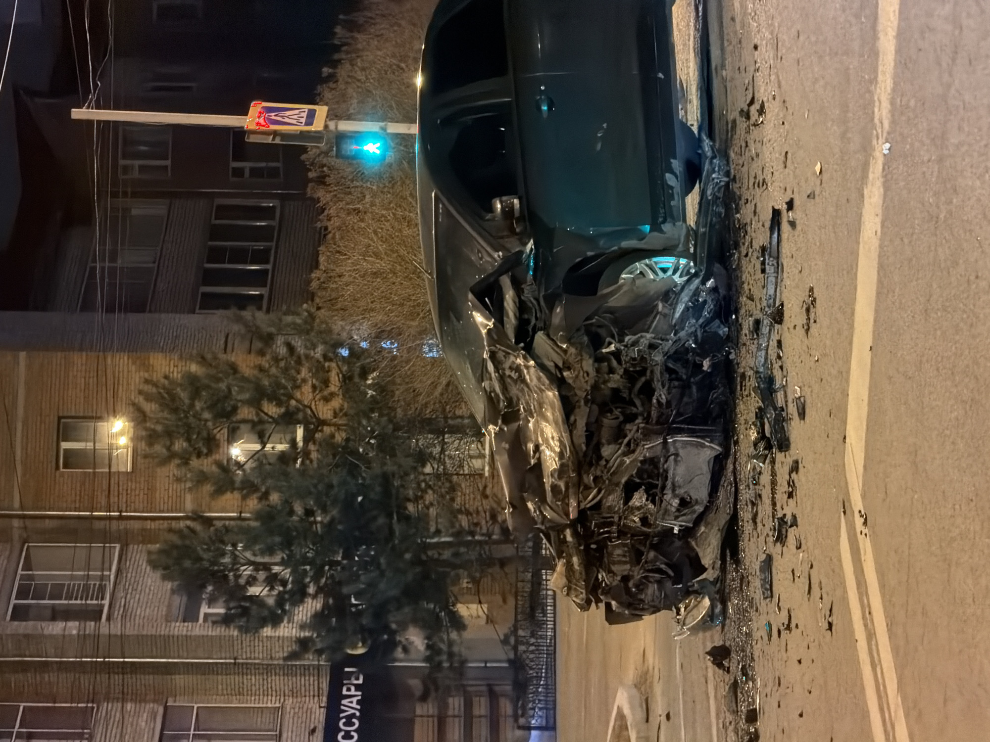 This media file captures the aftermath of a severe car accident at night in Krasnoyarsk, Russia.
**Subjects:** The primary subject is a dark-colored vehicle, likely an SUV or large sedan, with its front end completely destroyed and crumpled. The driver's side door is ajar. Surrounding the car on the asphalt road is a large amount of debris, including shattered plastic, metal fragments, and glass. In the background, there is a multi-story building with several illuminated windows. Bare trees are visible, some partially obscuring the building. Overhanging the street is a traffic light showing a green signal for what appears to be through traffic, next to a pedestrian crossing sign.
**Activity or event taking place:** The image depicts the scene following a serious vehicle collision. The extensive damage to the car's front suggests a high-impact crash.
**Notable details:**
* **Time of day:** It is night, as evidenced by the dark sky, artificial street lighting, and illuminated windows in the background building.
* **Weather:** The road appears dry, indicating no recent rain or snow.
* **Location cues:** The presence of a traffic light, pedestrian crossing sign, road markings (including a white line on the asphalt), and a residential or commercial building places the scene on a city street or intersection. The green traffic light is prominent. No people are visible in the immediate vicinity of the accident.
* **Condition:** The car is mangled beyond repair, with its engine compartment completely crushed and internal components exposed. Debris is spread widely across the road surface.
**Visible text:** A vertical street sign to the bottom left shows Cyrillic letters, partially reading "ССУАРЫ" (SSUARY), likely part of a word like "АКСЕССУАРЫ" (AKCESSUARY, meaning "accessories"). The pedestrian crossing sign has a blue background with a white pedestrian figure, and a red graphic (possibly a logo or additional symbol) superimposed on it.