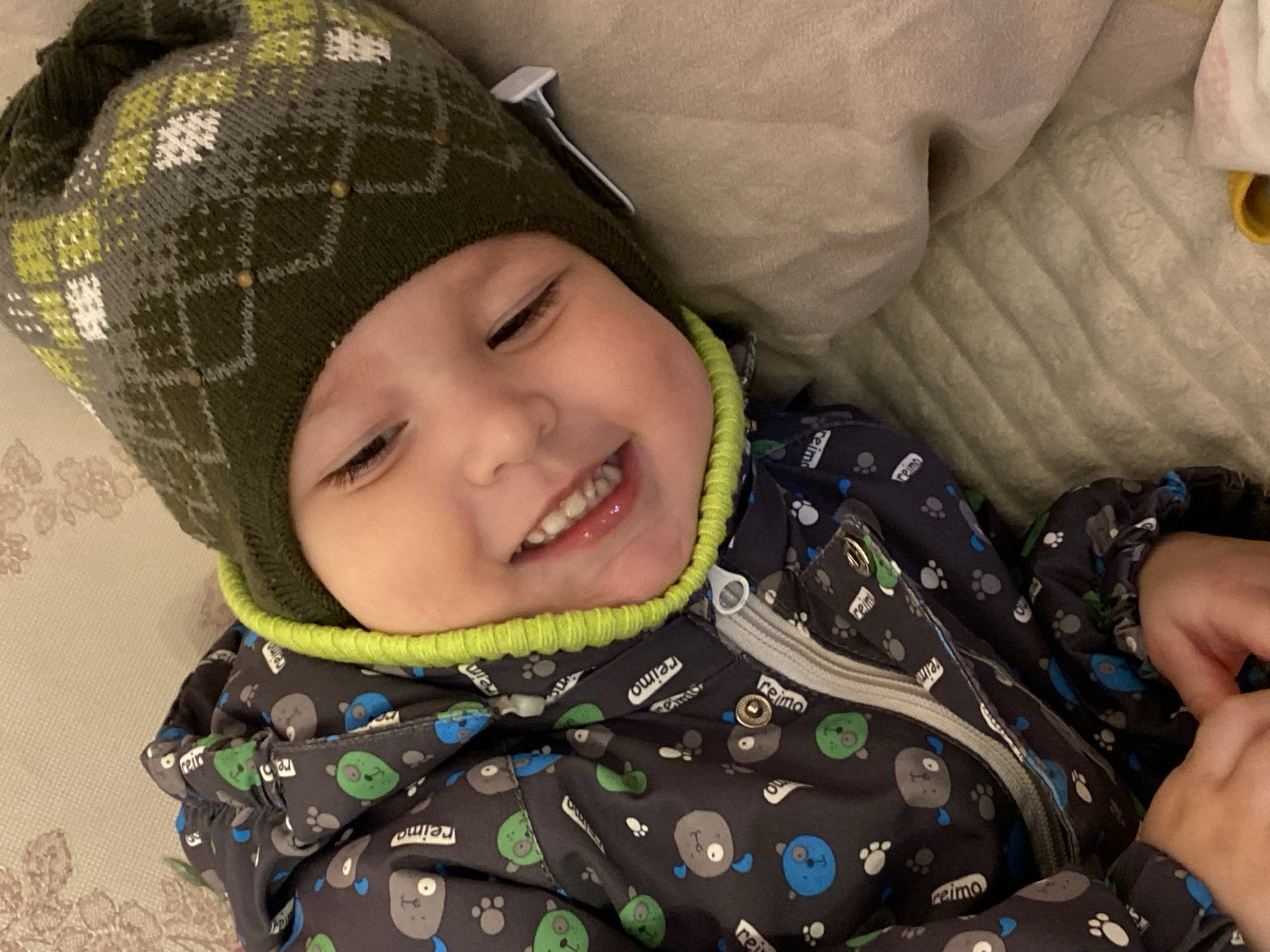 A young child is lying on a textured surface, possibly a bed or couch, wearing a patterned winter hat and jacket. The hat is predominantly dark green with gray, white, and light green diamond patterns. The jacket is dark-colored with a repeating pattern of cartoonish animal faces and paw prints. A bright green trim is visible around the neck of the jacket. The child is smiling with their teeth showing. The location is Марковский, Russia.