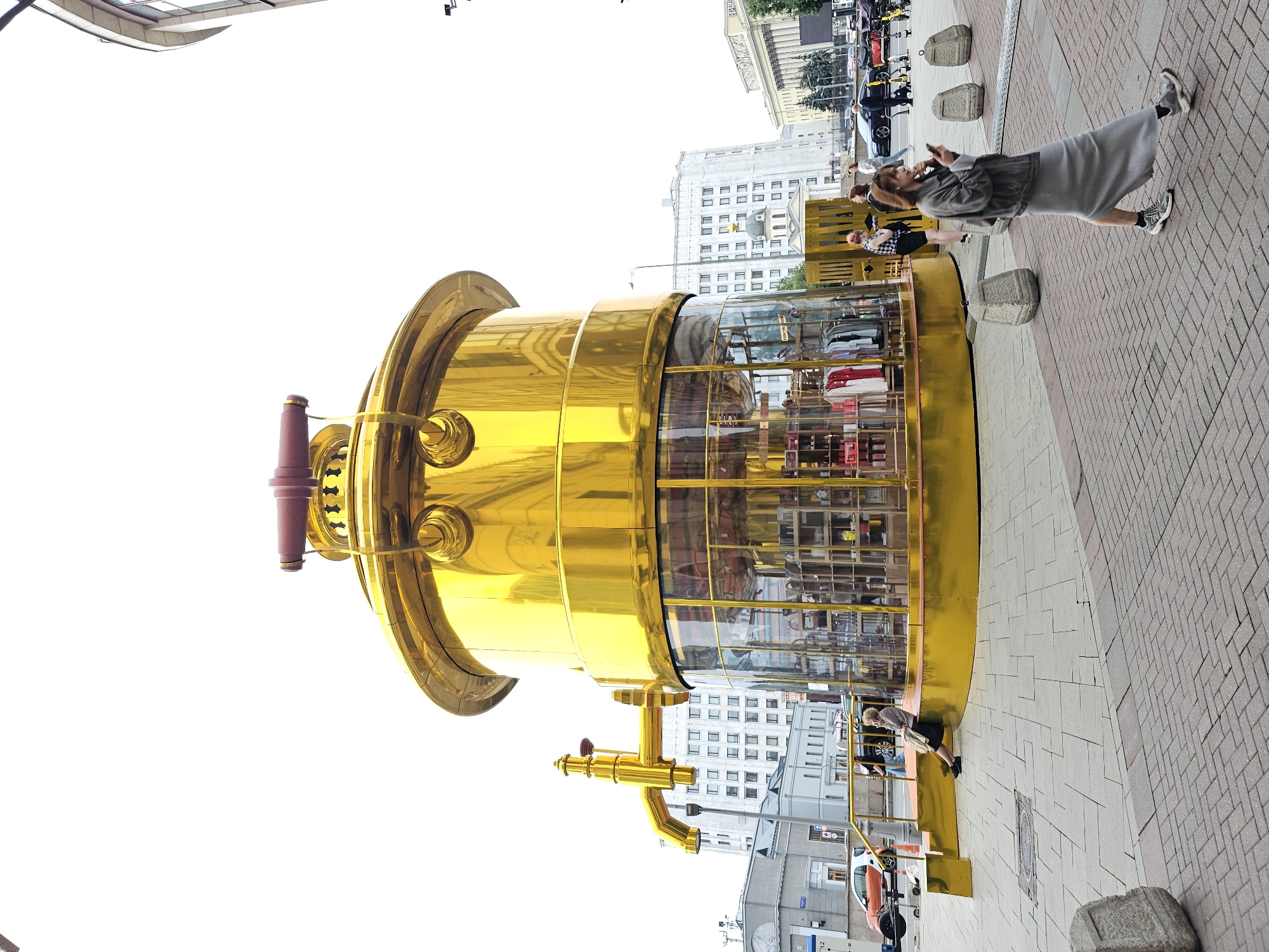 This is a vertical, outdoor, daytime photograph taken in Moscow, Russia. The main subject is a large, shiny, golden, cylindrical structure that appears to be a shop or a kiosk. It has a transparent glass section showcasing various items, possibly clothing, displayed on shelves. The structure is adorned with metallic accents and a dark red handle-like element on one side.
In front of the golden structure, several people are visible on a brick-paved sidewalk. A woman in a grey dress and jacket is looking at her phone. Another person in a black and white checkered shirt is seen leaning. Further back, other individuals are walking or standing. The background consists of tall, multi-story buildings with a classical architectural style, some featuring many windows. The sky is bright, suggesting fair weather. The scene appears to be a public urban space, possibly a pedestrian zone or a shopping street, with elements of city architecture and everyday street life.