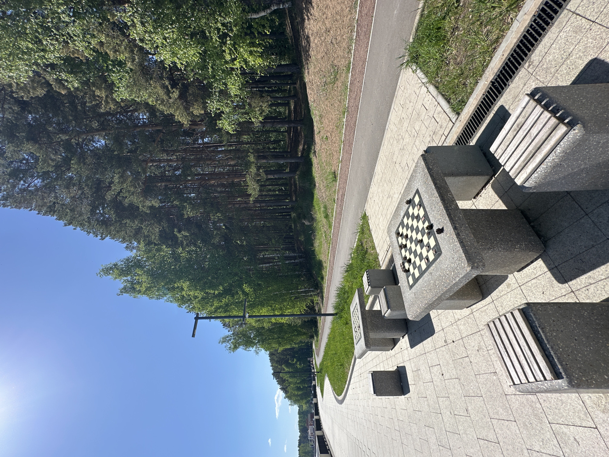 The image depicts a public space in the city of Chaikovsky, Russia, featuring a paved walkway and a concrete chess table with attached benches. The chess table has a black and white checkered board, and the benches have wooden slats. The walkway is bordered by grass and trees on one side and a low retaining wall on the other. A lamppost is visible along the walkway. The sky is clear and blue. The scene appears to be a park or recreational area.