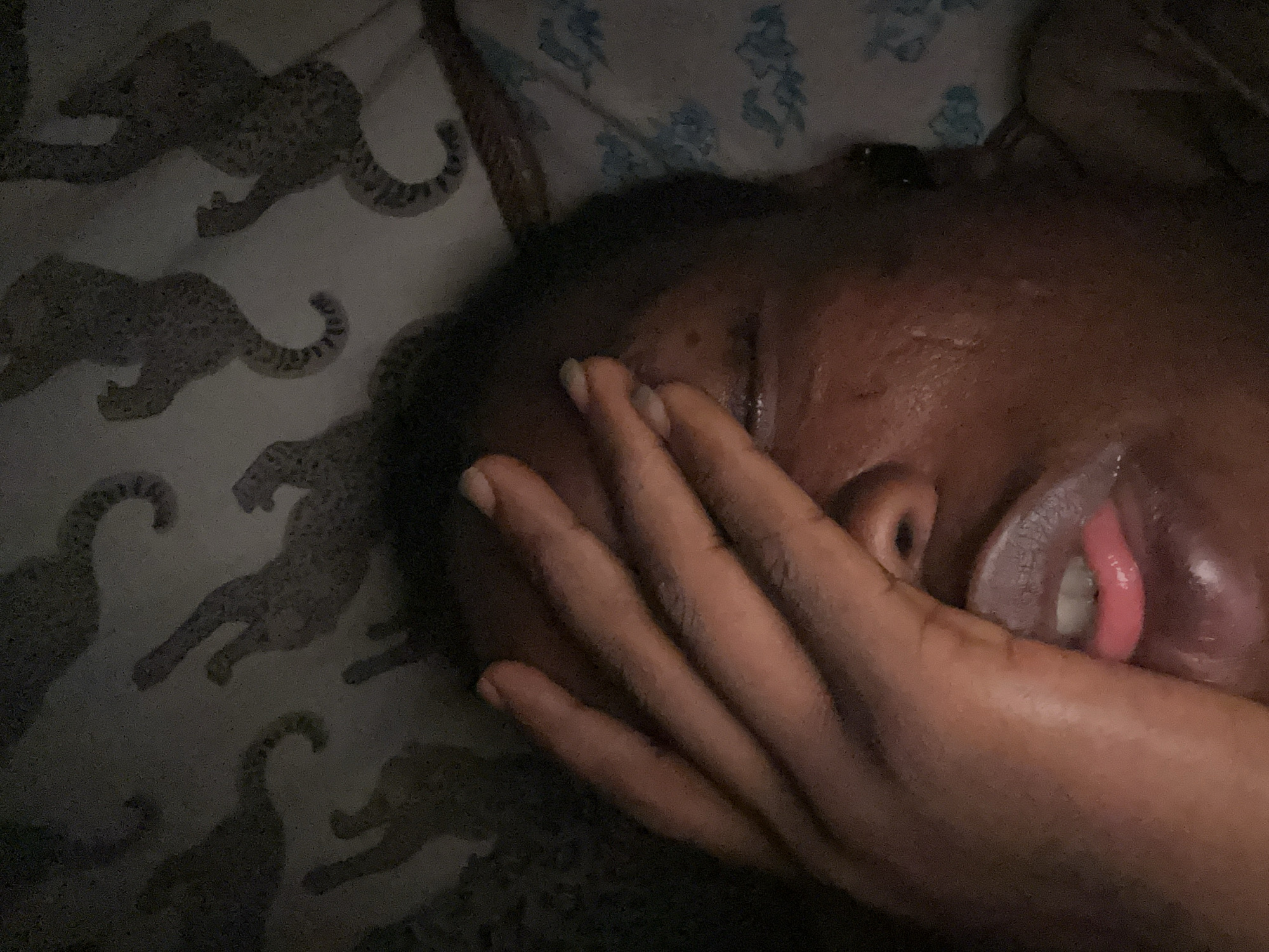 A close-up, slightly blurred, low-light photograph shows a person lying down, presumably in a bed. The person's right hand is raised to their face, partially obscuring it. Part of their nose, mouth, and a tooth are visible. The person has dark skin. The bedding has a leopard print pattern on the left and a blue floral pattern in the upper right. Location information is unavailable.
