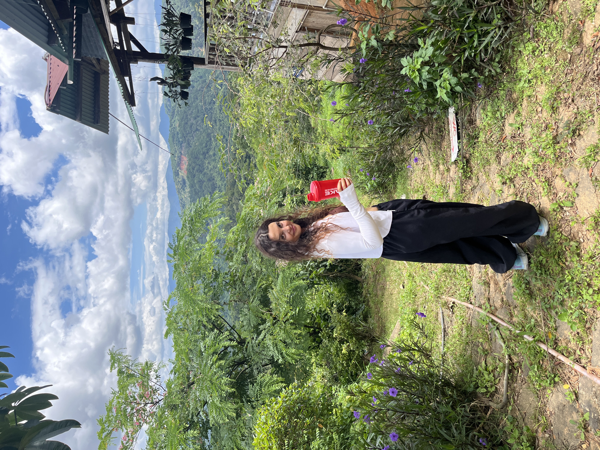 A young woman with curly brown hair stands in a lush, green outdoor environment with a view of rolling hills in the distance. She is wearing a white long-sleeved shirt and loose black pants, and holding a red shaker bottle in her right hand. The ground around her is covered in grass, stones, and some purple flowers. To her right, there is a rustic wooden structure with a corrugated metal roof and hanging plants. The sky is partly cloudy with a bright blue expanse. The location is the city of Sơn Điền, Vietnam.