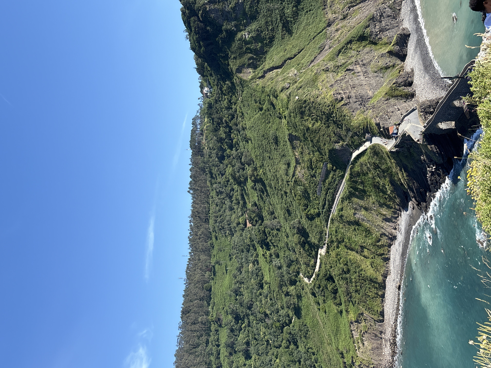 The image depicts the ascent to the hermitage of San Juan de Gaztelugatxe, located in the municipality of Bakio, Spain. A winding paved road with white railings snakes its way up the lush green hillside, with several people visible walking along it. The road connects to a stone bridge that spans the sea, leading to the rocky promontory where the hermitage is situated. The coastline features rocky beaches and the turquoise water of the sea crashes against the shore. The sky above is clear blue with a few wispy clouds. A house is visible nestled on the hillside in the distance.