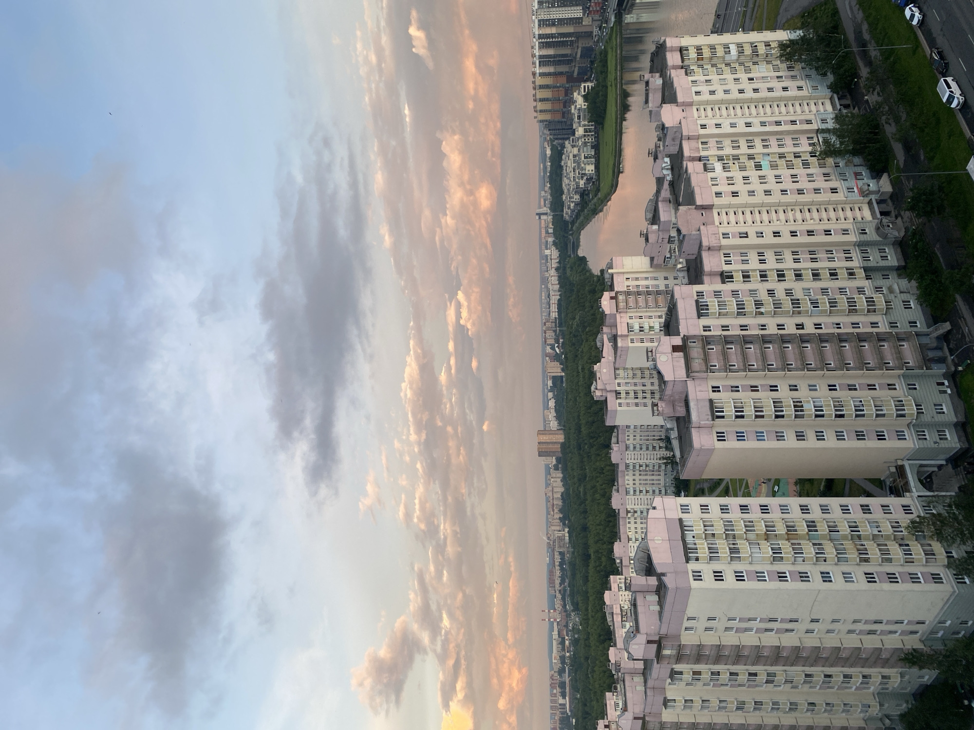 The image depicts a panoramic view of the city of Saint Petersburg, Russia, during sunset. The sky is filled with soft, pastel-colored clouds, transitioning from light blue to hues of pink and orange. In the foreground, several tall residential buildings with light-colored facades and pink accents are visible. These buildings appear to be part of a modern housing complex.
Beyond the immediate residential area, a wide river or canal cuts through the urban landscape, reflecting the colors of the sky. A dense green forest stretches along the riverbanks, providing a natural buffer within the city. Further in the distance, a mix of industrial structures, including smokestacks, and other residential and commercial buildings are scattered across the horizon. The overall impression is one of a vast urban area situated alongside a natural waterway and a significant green space. No people or specific actions are discernible in the image.