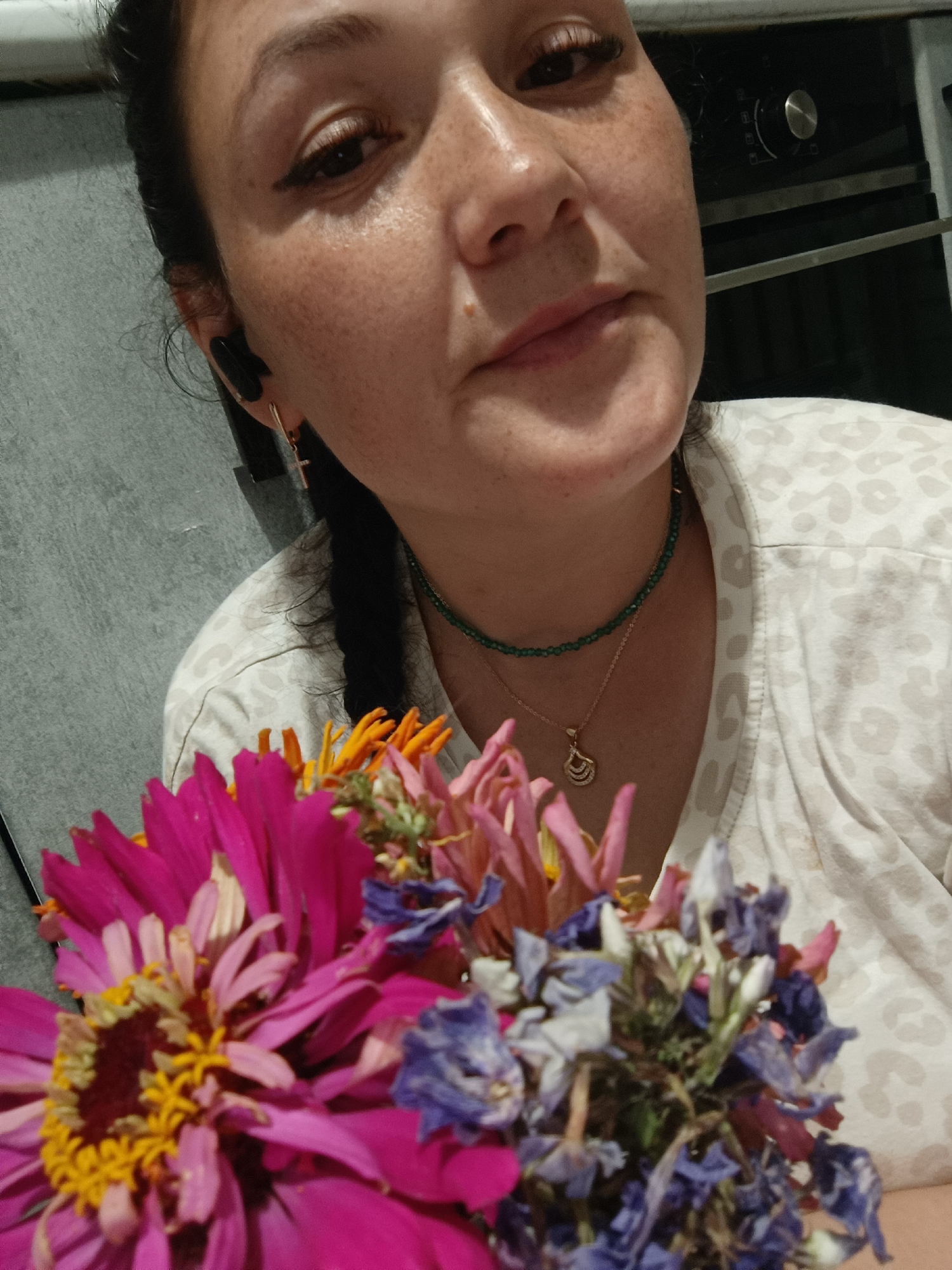 In the city of Chaikovsky, Russia, a close-up, medium-shot photograph captures a woman holding a bouquet of flowers. The woman, with dark hair and freckles across her nose and cheeks, is looking slightly upward and to her right. She is wearing black earbuds, a gold cross earring in her left ear, a green beaded necklace, and a gold pendant. Her top has a light-colored leopard print pattern. The bouquet in the foreground consists of a vibrant pink zinnia, along with dried flowers in shades of purple and orange. The background is blurred, showing a gray textured wall and what appears to be kitchen appliances. The lighting suggests an indoor setting at night.