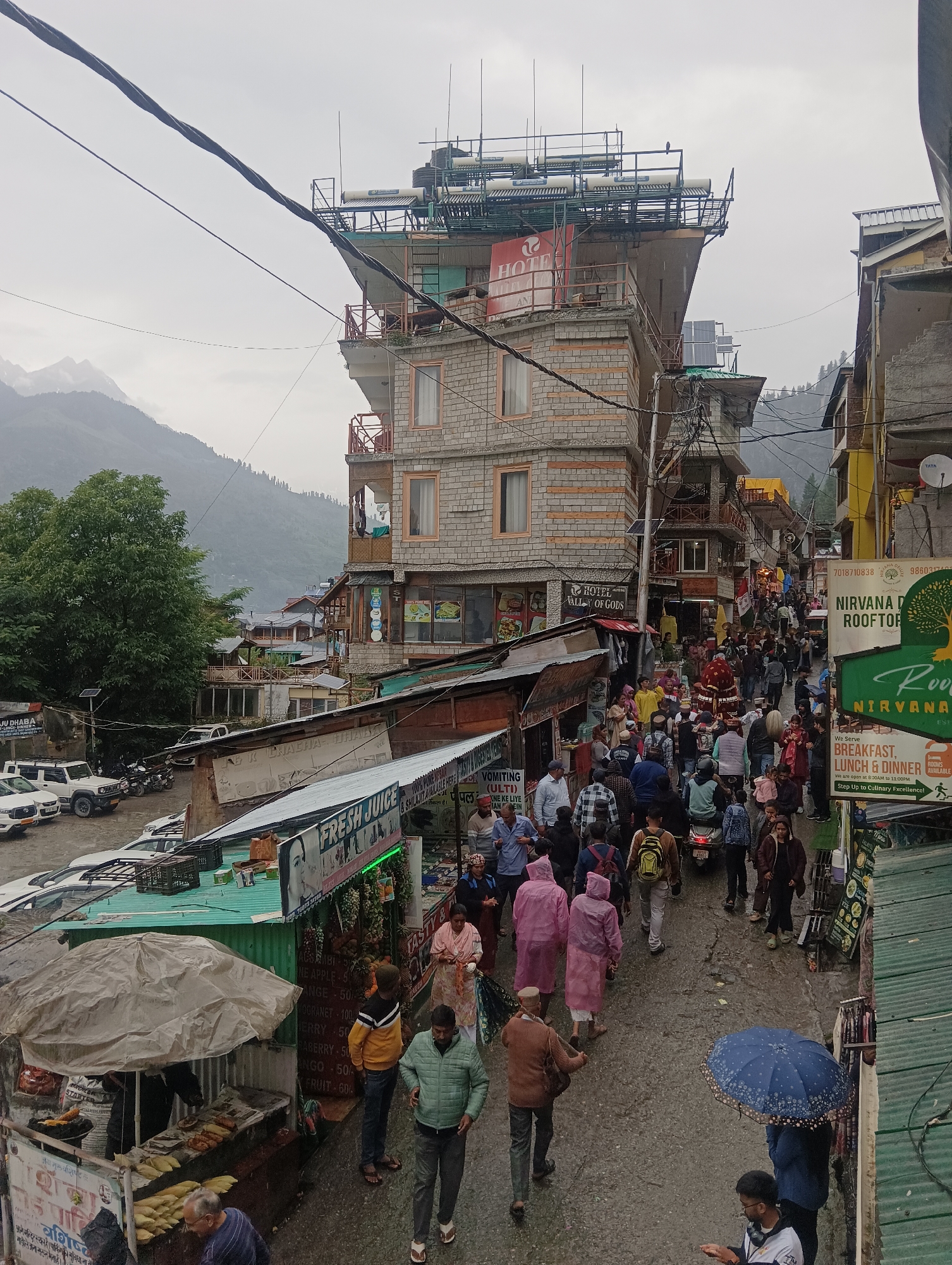 The image depicts a crowded street scene in Vashisht, India, under an overcast sky. Buildings, several stories high, line both sides of the narrow, wet street. Signs indicate businesses such as "Hotel Valley of Gods," "Nirvana Rooftop," and "Fresh Juice." People are walking in both directions, some under umbrellas, suggesting rain. Several individuals are wearing brightly colored raincoats. Vendors appear to be selling food and drinks from stalls along the street. In the background, mountains are visible, partially obscured by fog or clouds. A few parked vehicles are present on the left side of the street. The overall atmosphere is one of a bustling marketplace during inclement weather.
