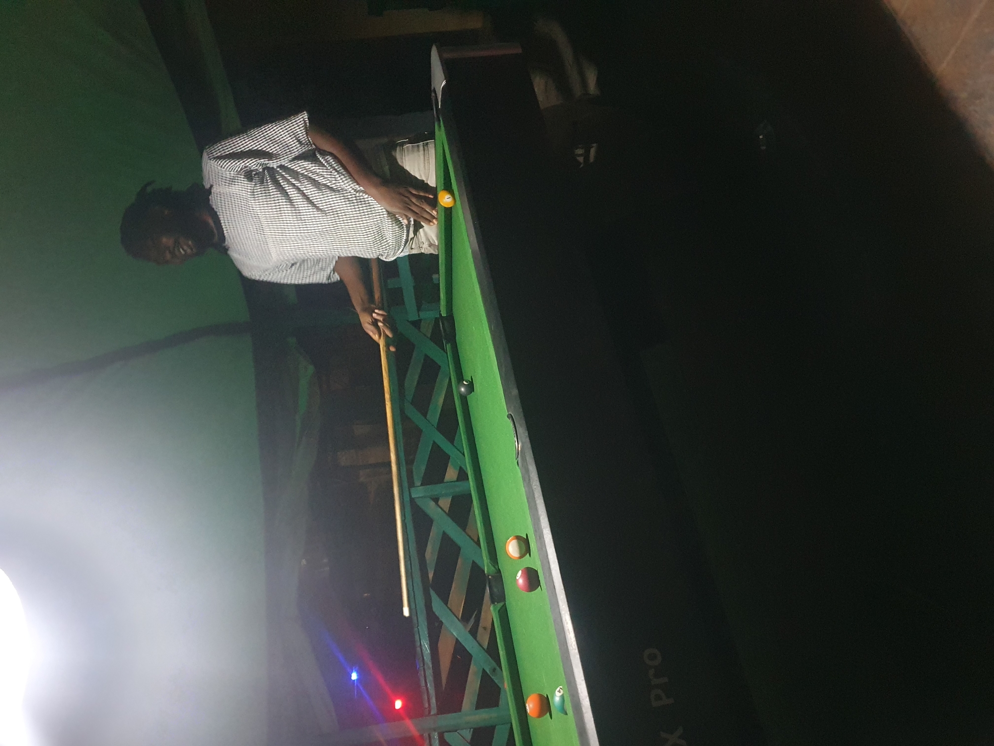 A full shot shows a Black man playing pool. He is standing at a pool table that has a green felt surface and wooden rails. There are several billiard balls on the table, including an orange ball, a red ball, a striped yellow and white ball, and a solid yellow ball with the number "1" on it. The man is wearing a black and white checkered t-shirt and light-colored shorts. He is holding a pool cue in his hands, and he is looking down at the table. The background of the image is dark, but there is a green structure behind the pool table that appears to be a railing or fence. There are also some lights in the background, including a blue and red light. The overall setting appears to be an outdoor area, possibly a bar or recreational facility. The image's metadata indicates the location is Lokoja, Nigeria.