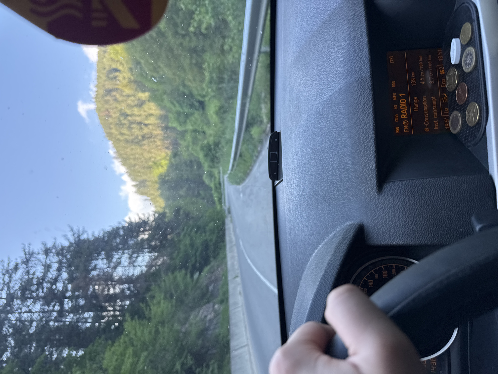 A forensic examination of the image reveals a first-person perspective from inside a vehicle, capturing a winding mountainous road in Sokolovtsi, Bulgaria. The asphalt road curves to the right, bordered by a concrete barrier on the left and a metal guardrail on the right. Dense green foliage and tall trees cover the steep slopes ascending on both sides, with a clear blue sky and sparse white clouds overhead. Sunlight illuminates the distant hills.

Within the vehicle, a human hand is visible on the dark, textured steering wheel, with a partial view of the instrument cluster showing a speedometer marked up to 200 km/h and a digital time display of "14:36". The central console features a digital display showing "FM RADIO 1", a "Range" of "139 km", an "Ø-Consumption" of "4.5 Lt./100 km", and "Inst. consumpt" of "8.7 Lt./100 km", alongside "19.5° Lo Eco 2 18:51". Below this display, a small compartment holds several Euro coins (including 1 and 2 Euro denominations) and one white, circular tablet. A dark, rectangular sensor is affixed to the dashboard near the base of the windshield. A portion of a brown and red sticker is visible in the upper right corner of the windshield. The windshield itself shows minor dust or debris.