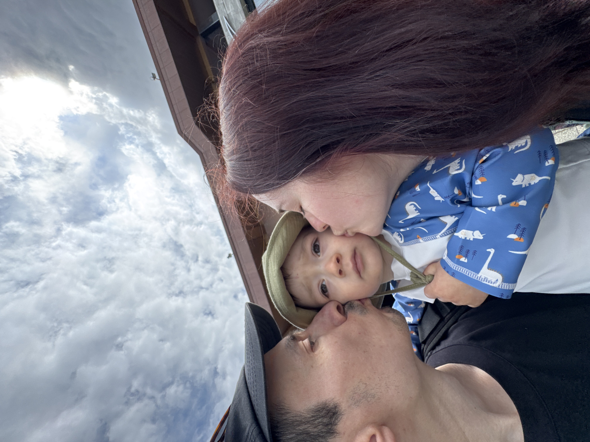 A close-up, tilted photograph captures a family moment against a backdrop of a cloudy sky and a partial view of a building. A woman with dark auburn hair is positioned on the right side of the frame, kissing a young child on the forehead. The child, wearing a light green sun hat and a blue patterned shirt with white long sleeves, is held in front of the woman. A man wearing a black baseball cap is located at the bottom of the frame, kissing the child’s cheek. The sky, filled with gray and white clouds, occupies the upper left portion of the image. The building structure, likely part of a roof or awning, is visible in the upper middle. The setting is presumed to be Sochi, Russia, based on location instructions, though no explicit landmarks are visible in the image.