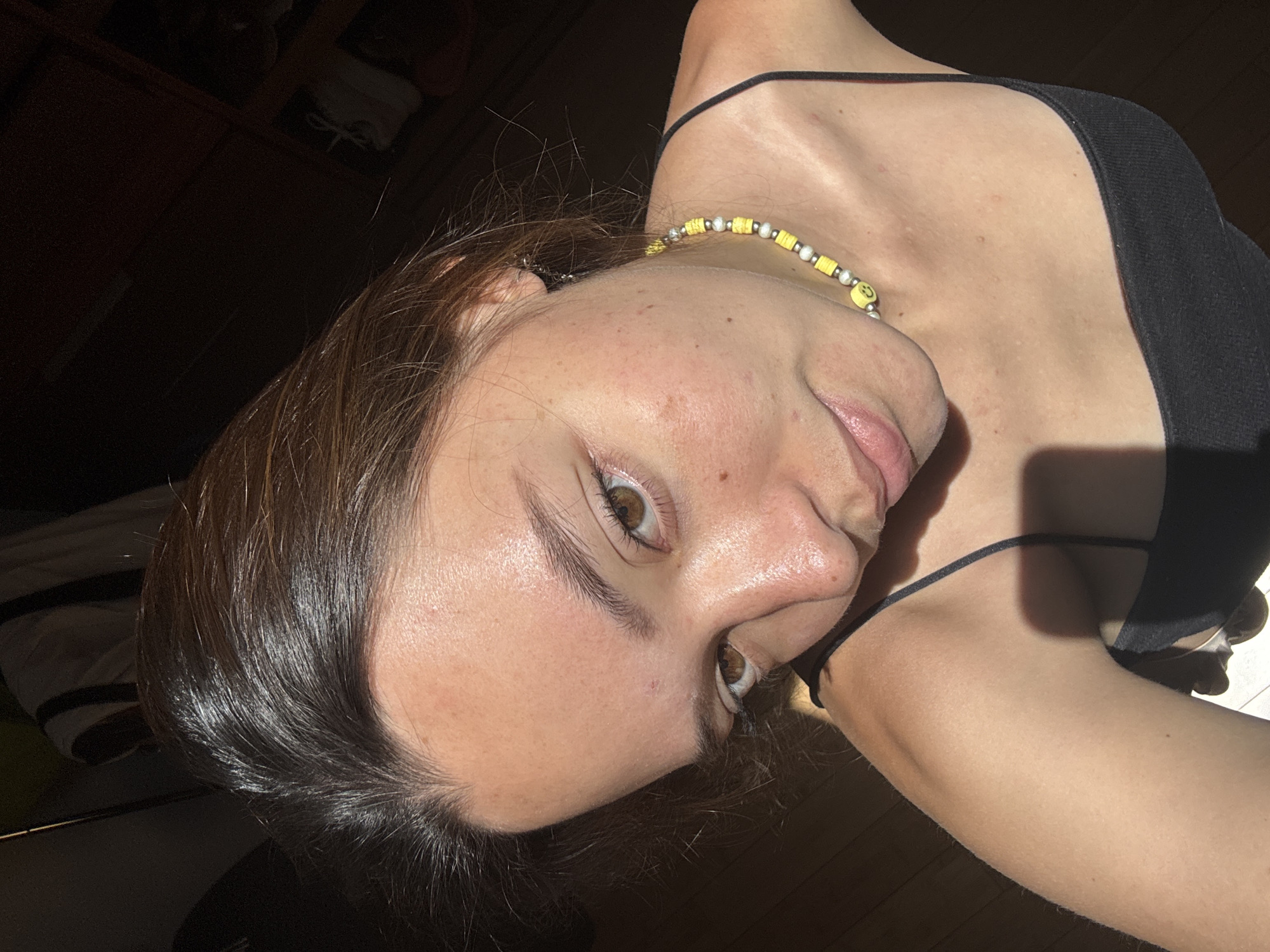 A close-up portrait of a young woman is captured indoors in San Bernardino Tlaxcalancingo, Mexico. The image is taken from a slightly high angle and rotated counter-clockwise. The subject, likely in her late teens or early twenties, has brown hair, fair skin with some freckles, and light brown eyes. Her gaze is directed towards the viewer, with a neutral to slightly gentle expression.
She is wearing a black tank top with thin spaghetti straps. Around her neck is a beaded necklace composed of yellow, white, and silver beads, featuring a distinct yellow smiley face bead. Strong, directional sunlight illuminates the left side of her face, neck, and shoulder, creating bright highlights on her skin and hair, and casting shadows that define her features. This suggests the photo was taken during the day, possibly morning or late afternoon, with light entering from a window.
The background is largely dark and out of focus, revealing hints of indistinct objects or furniture in deep shadow, indicating an indoor environment. There is no other visible text apart from the smiley face on the necklace.